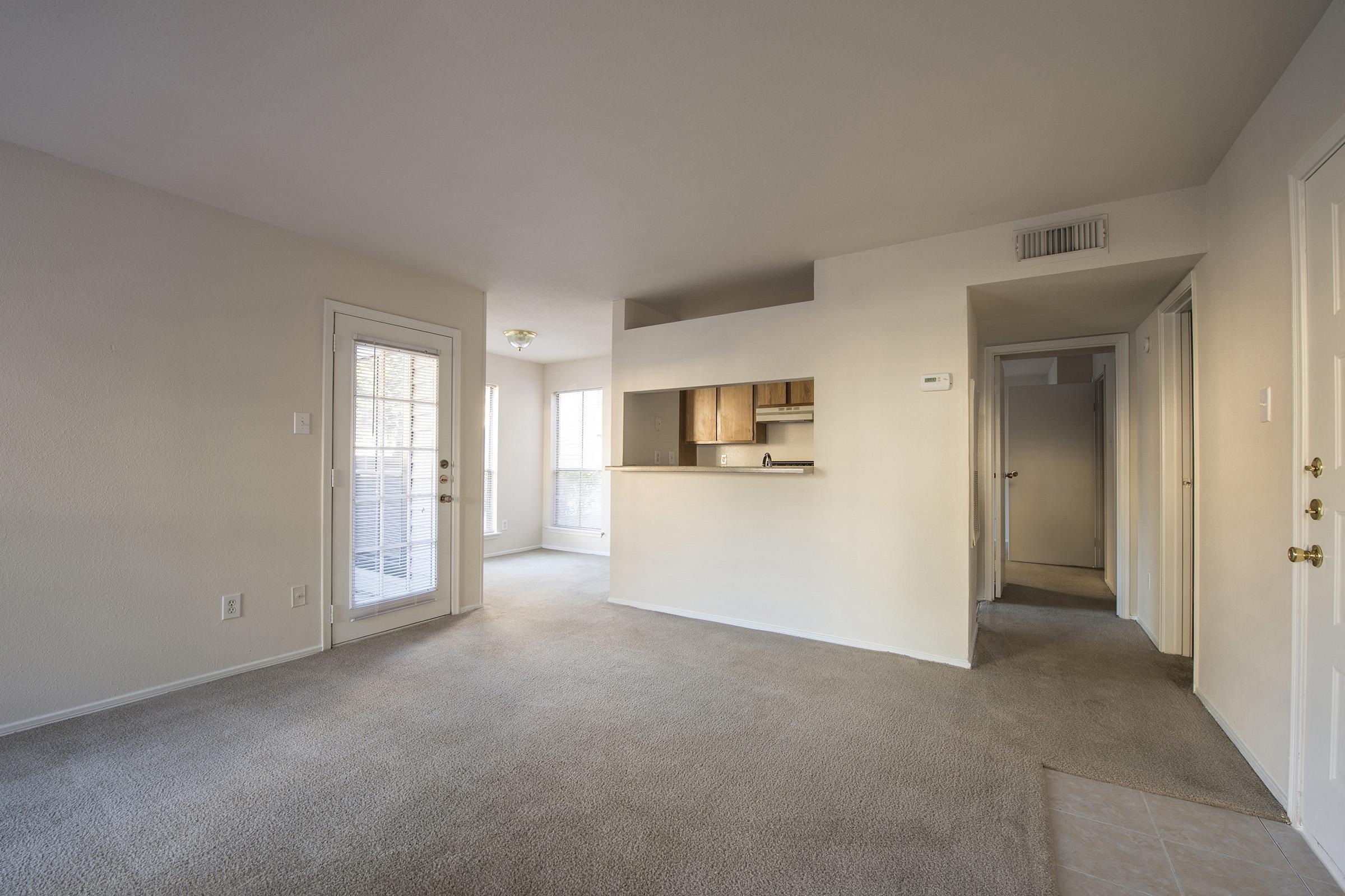 A spacious, empty living area with light-colored walls and carpet. On the left, there's a sliding glass door leading to a balcony. The kitchen area is visible at the far end, with wooden cabinetry. A hallway is seen on the right, leading to other rooms. Natural light comes in through multiple windows.