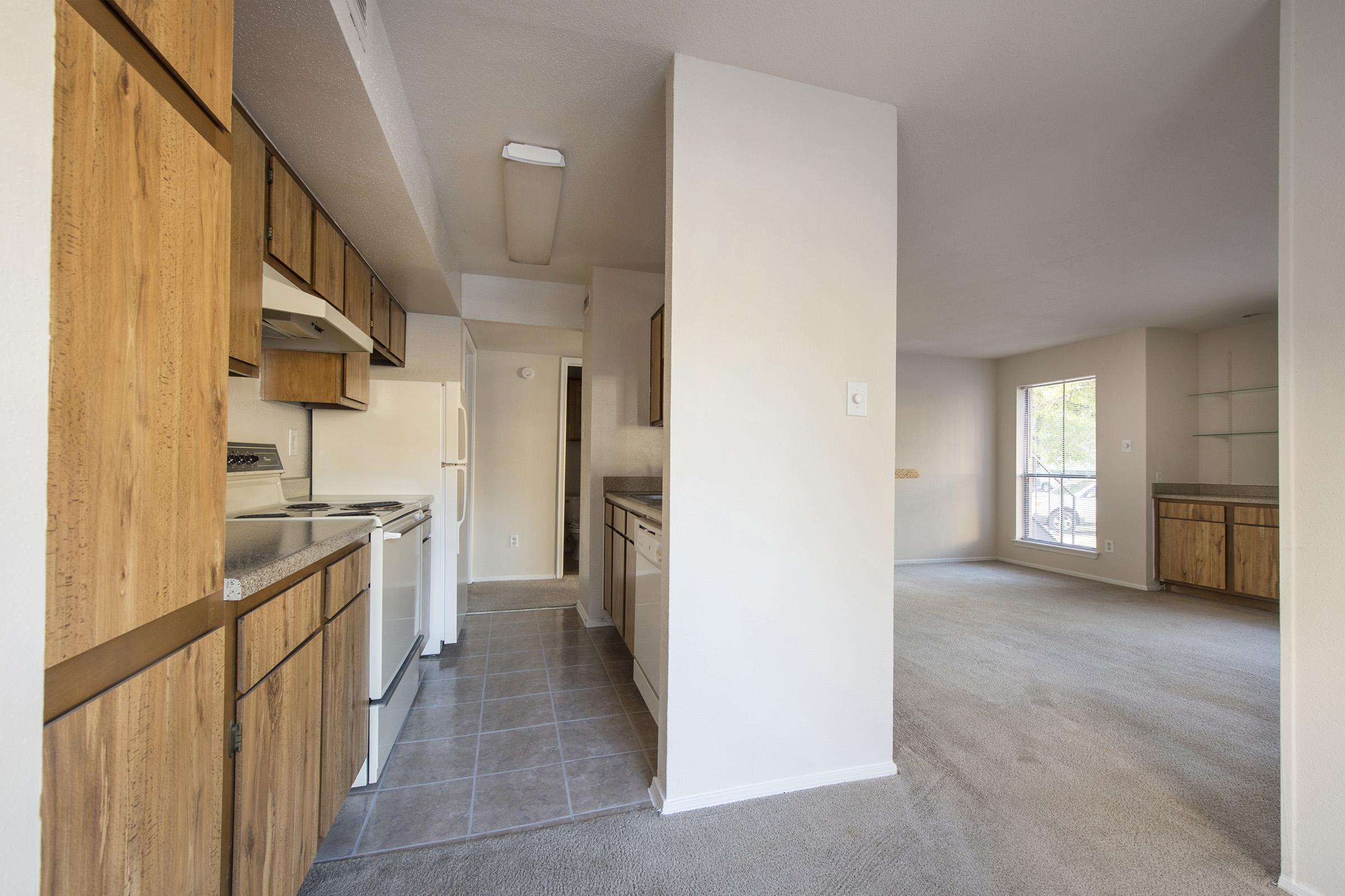 A view of an apartment interior featuring a kitchen on the left with wooden cabinetry, a white refrigerator, and an oven. The hallway leads to a spacious living area with light-colored carpet and large windows providing natural light. Shelving is visible in the room on the right.