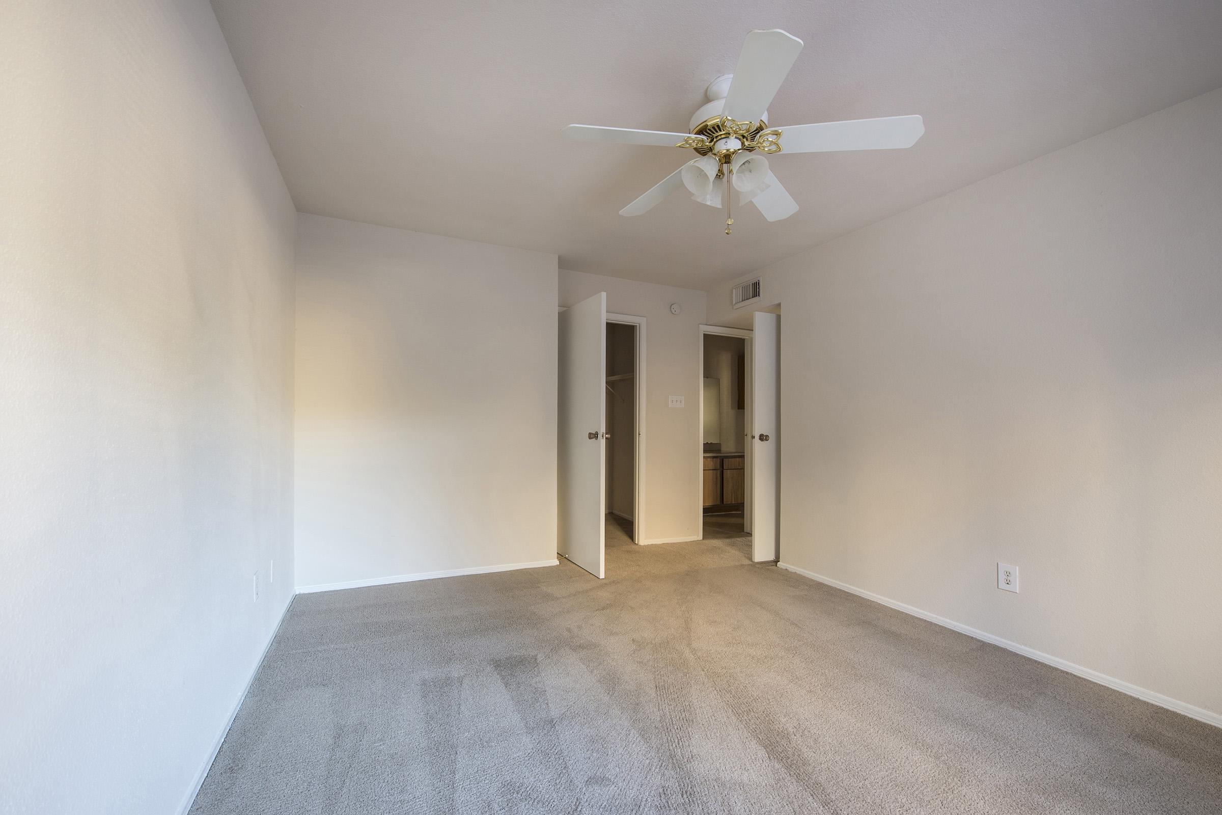 A spacious, vacant room with light beige walls and tan carpeting. A ceiling fan with three blades is installed in the center. Two open doors lead to adjoining rooms, and natural light partially illuminates the space, creating a calm and inviting atmosphere.