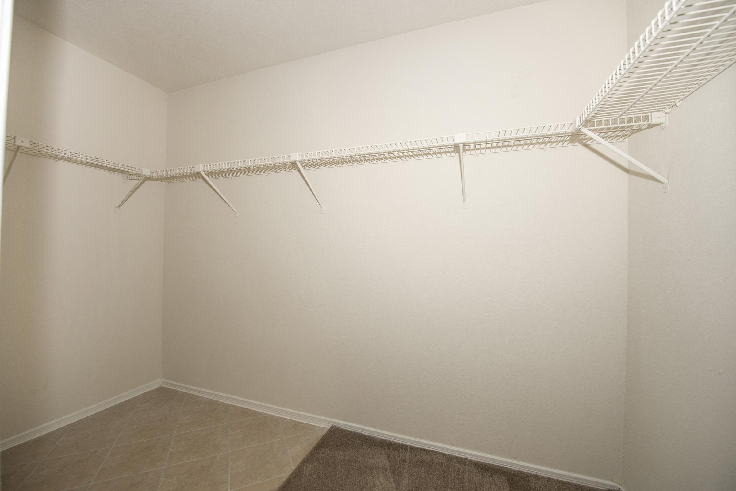 Empty closet with light-colored walls and a tiled floor. There are white wire shelves attached to the walls, providing storage space, but no items are currently stored on the shelves or the floor. The overall appearance is clean and minimalistic.