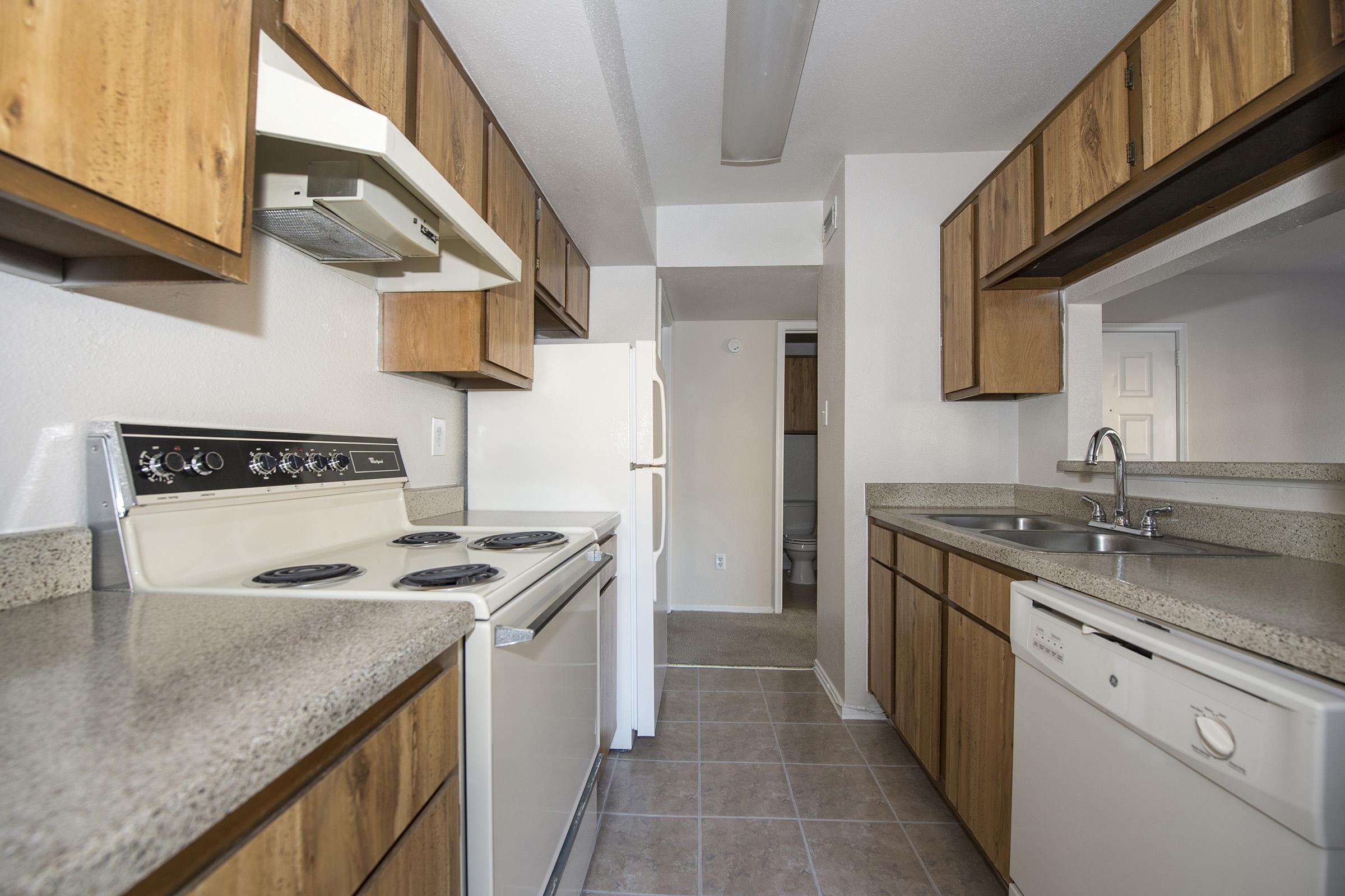 A modern kitchen featuring wooden cabinets, a white refrigerator, an oven with stovetop, a sink, and a dishwasher. The countertop is light gray, and the floor has beige tiles. The layout includes an open space with a view towards a nearby room.