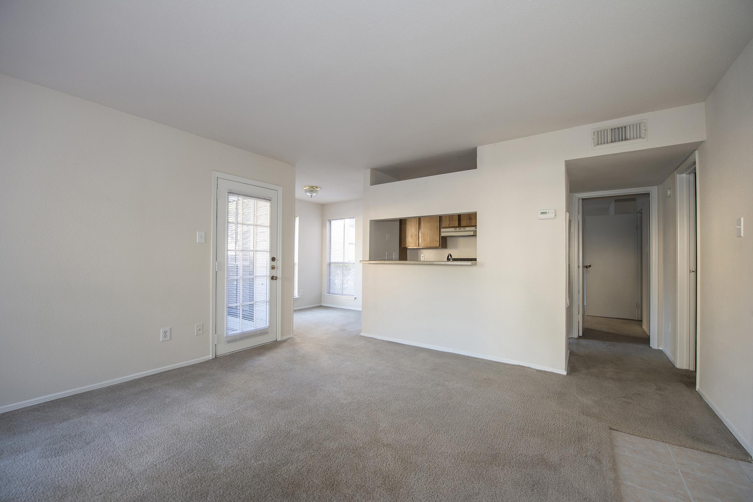 Interior of a spacious, unfurnished apartment with light beige walls and carpet. Features a small kitchen area visible in the background, large windows letting in natural light, and a sliding glass door leading to a balcony. A hallway extends to the right, leading to other rooms. Overall, bright and open layout.