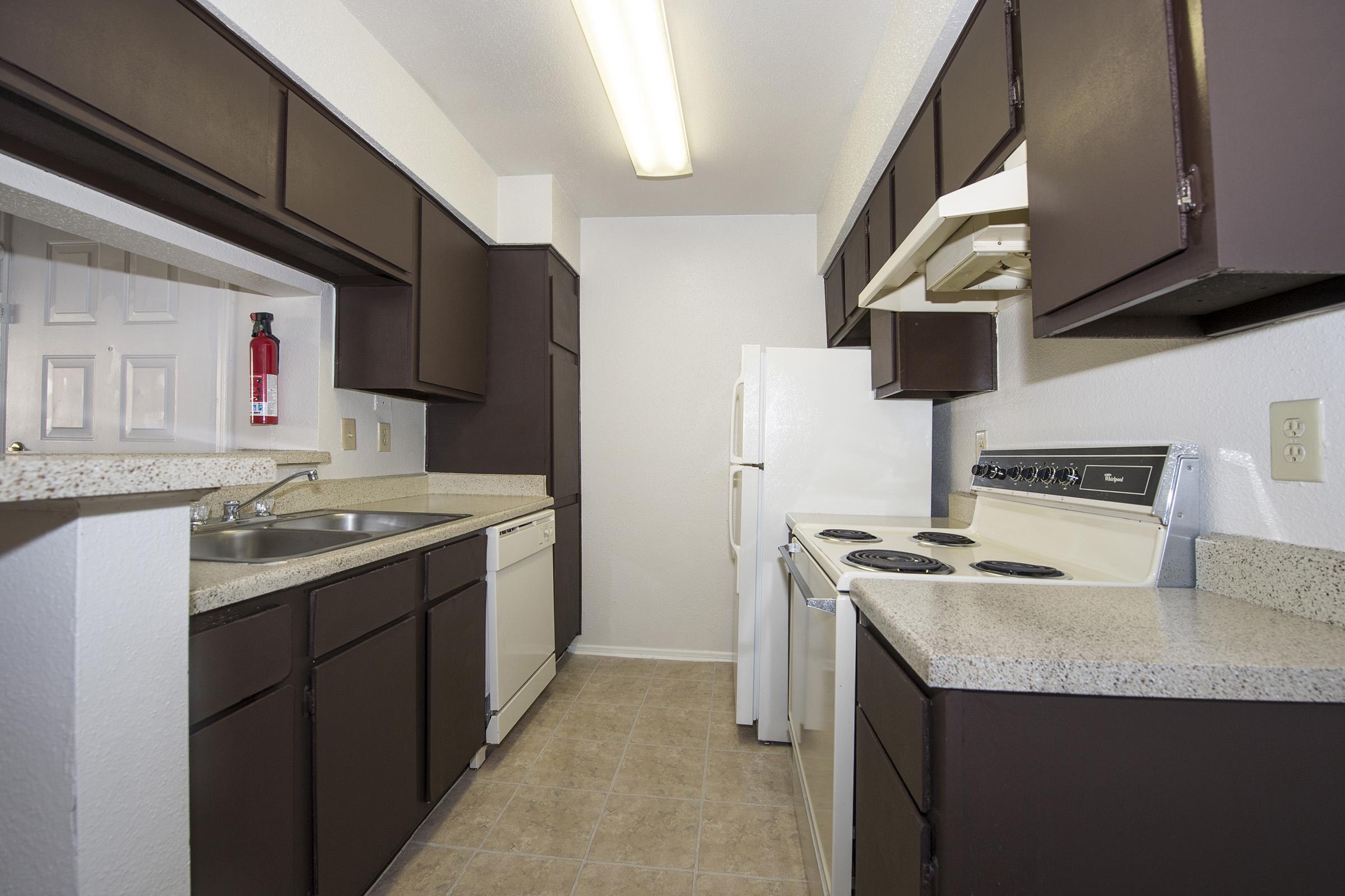 A small kitchen featuring dark brown cabinetry, a white refrigerator, a white stove with an oven, and a dishwasher. The countertops are light-colored, and there's a window providing natural light. The flooring is tiled in a neutral shade, and a fire extinguisher is mounted on the wall.