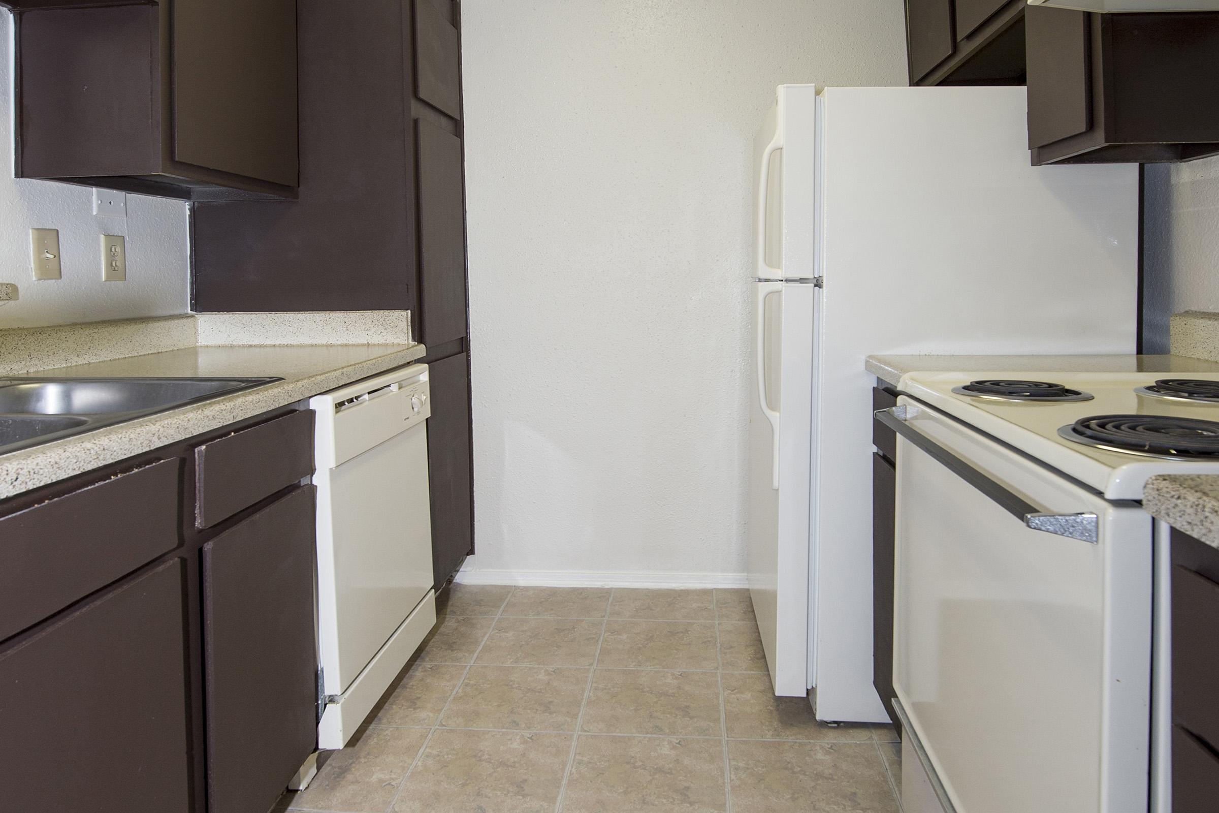 A modern kitchen featuring dark brown cabinetry, a white refrigerator, a white dishwasher, and a white stove with black burners. The countertops are light-colored, and the floor has beige tiles. The walls are painted a light shade, providing a clean and bright appearance.