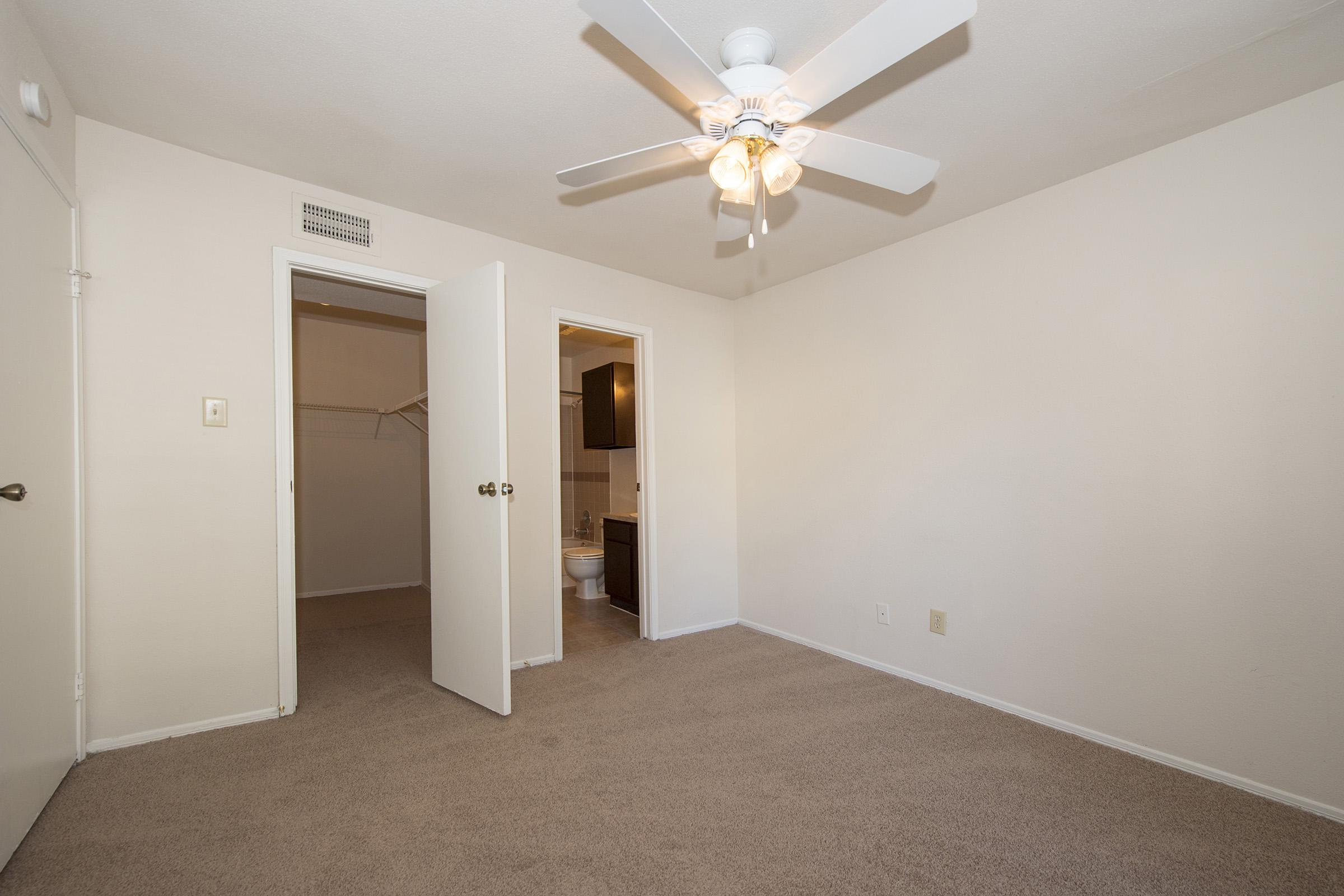 A clean, empty bedroom with light-colored walls and carpet. There is a ceiling fan with five light fixtures. Two open doors lead to a closet and a bathroom. The overall atmosphere is bright and spacious, suitable for various furnishings and decoration options.