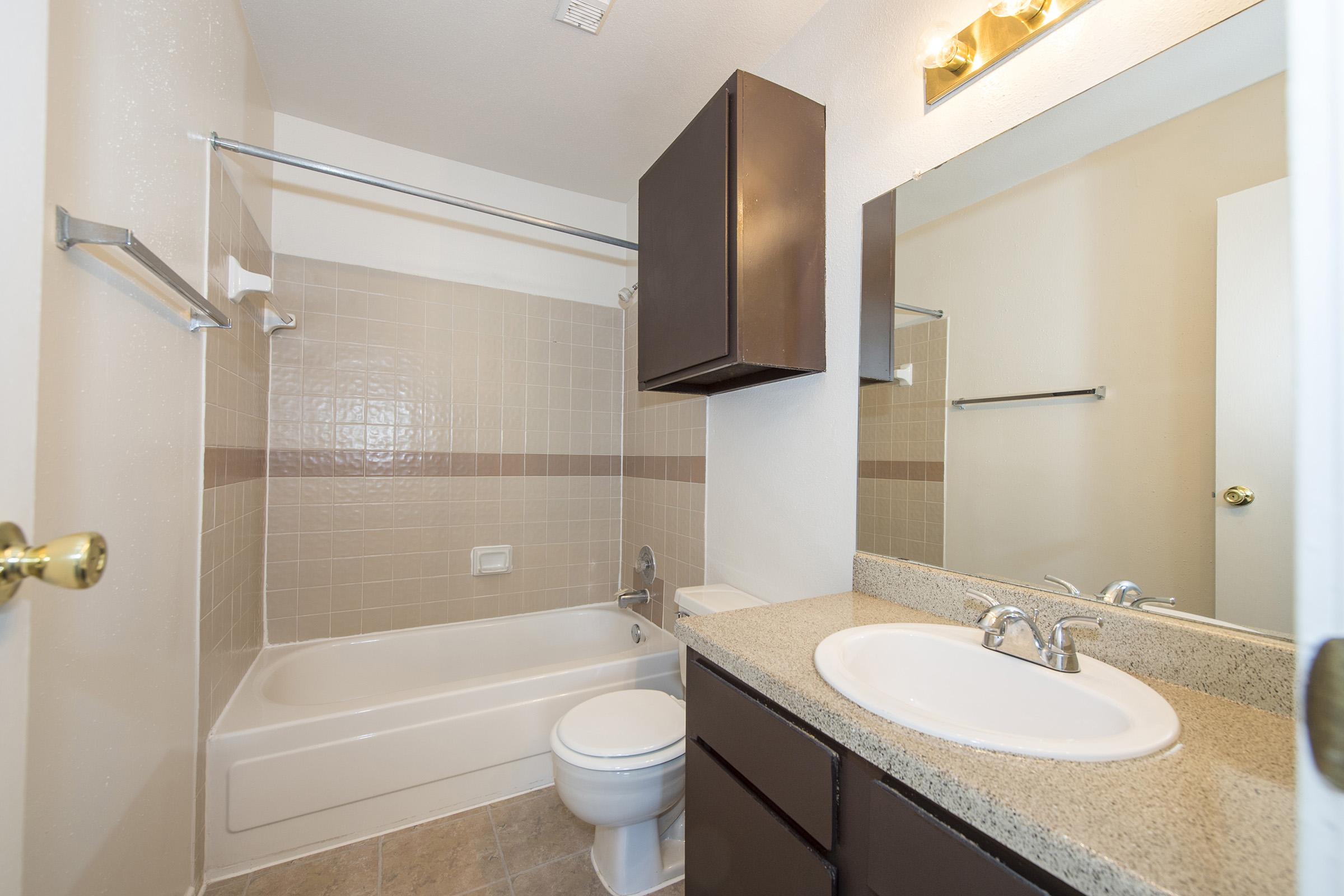 A clean, modern bathroom featuring a bathtub with a shower, beige tile walls, dark wood cabinets, a countertop sink, a large mirror, and bright lighting. The space is well-organized with essential bathroom fixtures and a neutral color scheme.