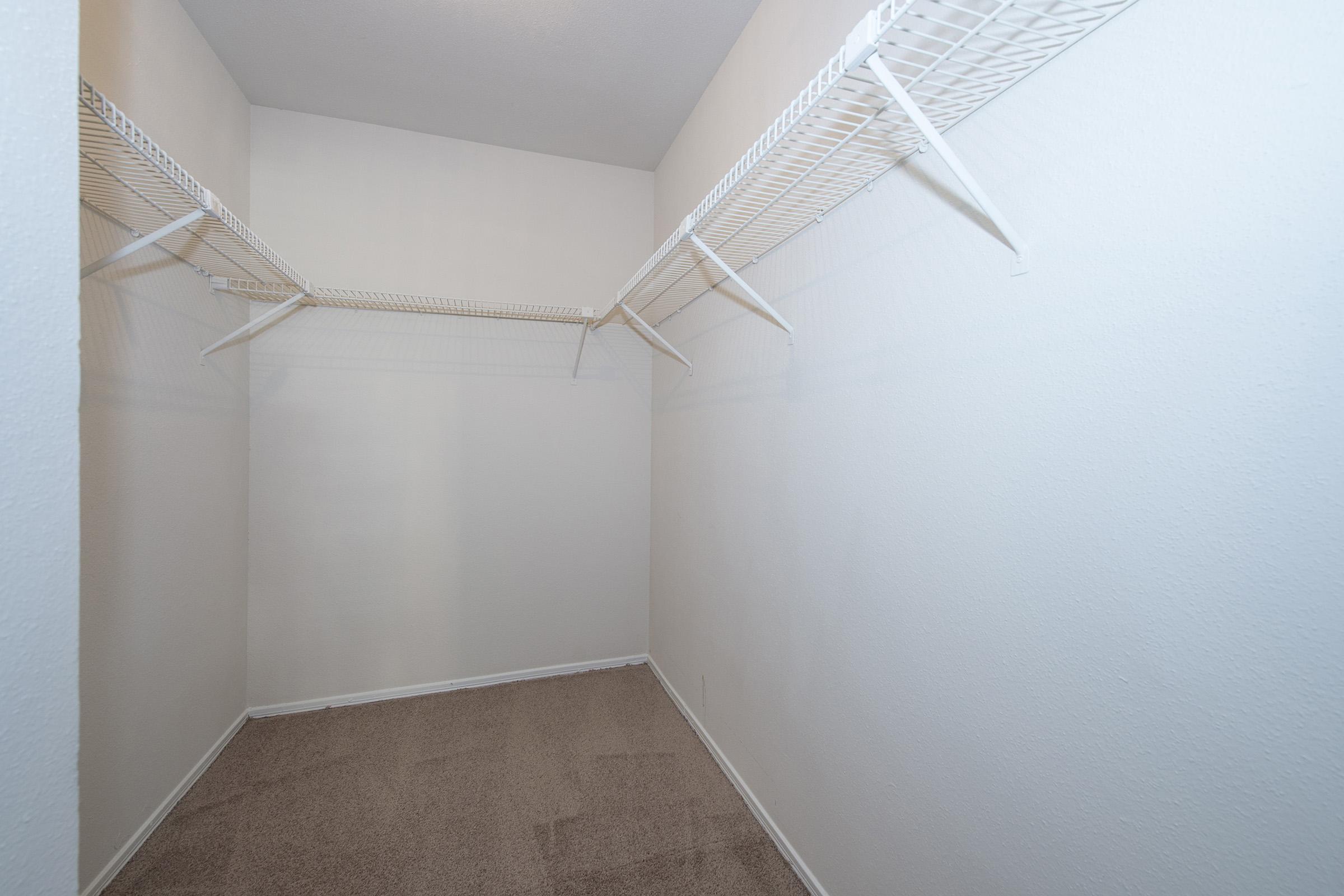 A clean, empty closet featuring white wire shelves. The walls are painted in a light color, and the floor is covered in beige carpet. The space is well-lit, with a simple, uncluttered design, suitable for organization and storage.