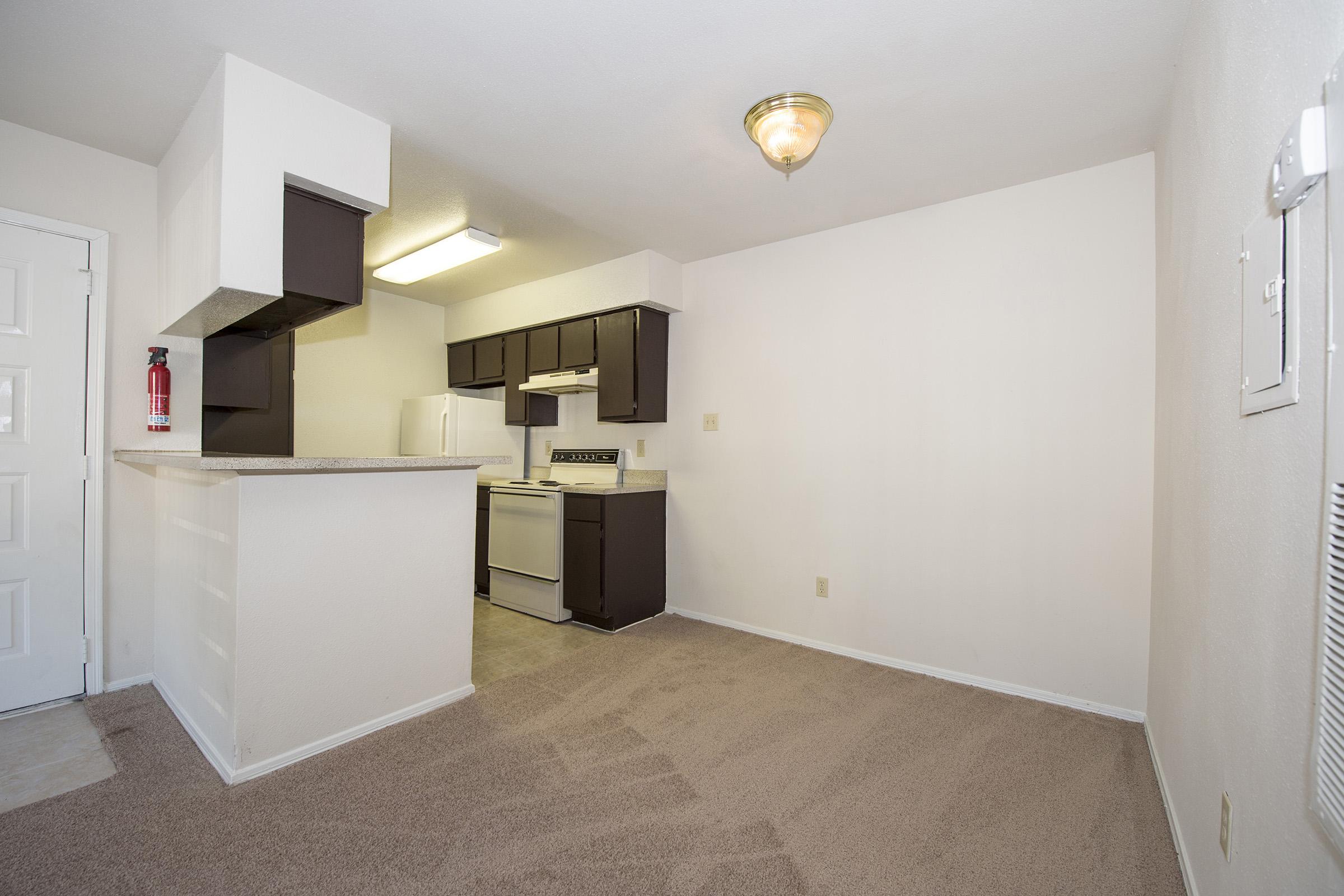 A small, well-lit kitchen and living area featuring beige walls and carpet. The kitchen includes dark brown cabinets, a white refrigerator, a stove, and a countertop with a small breakfast bar. A door leads out of the space, and there is a fire extinguisher mounted on the wall.