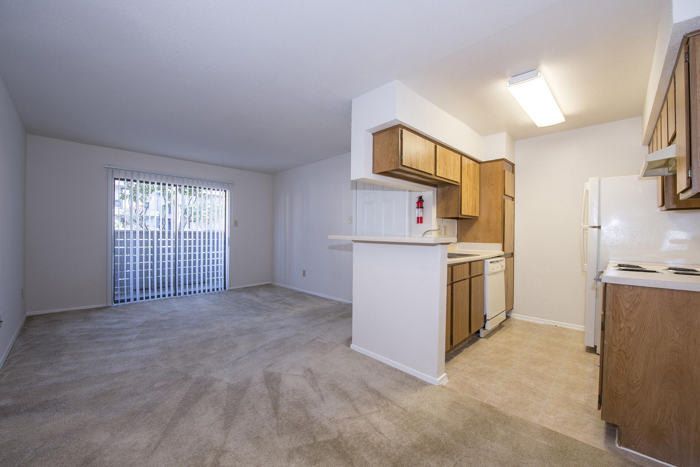 A spacious, unfurnished living area featuring beige carpeting, a small kitchen with wooden cabinets, and a white refrigerator. A sliding glass door leads to a balcony or patio area, while the walls are painted in a light color, enhancing the bright and airy feel of the space.