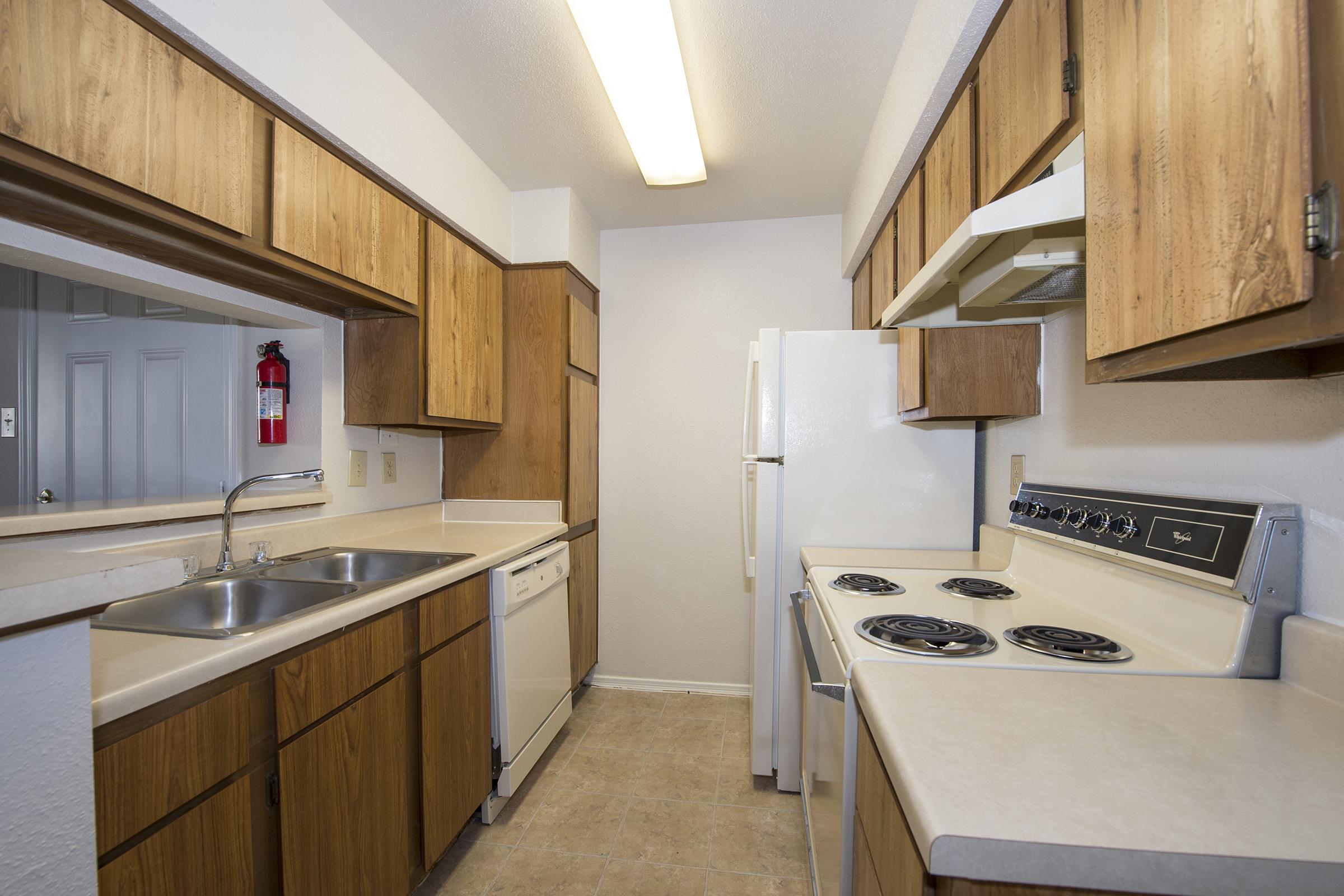 A modern kitchen featuring wooden cabinets, a double sink, a dishwasher, an electric stove, and a refrigerator. The space is well-lit with overhead lighting, and the walls are painted in a light color, creating a clean and inviting atmosphere. The floor is covered with a neutral-toned vinyl.