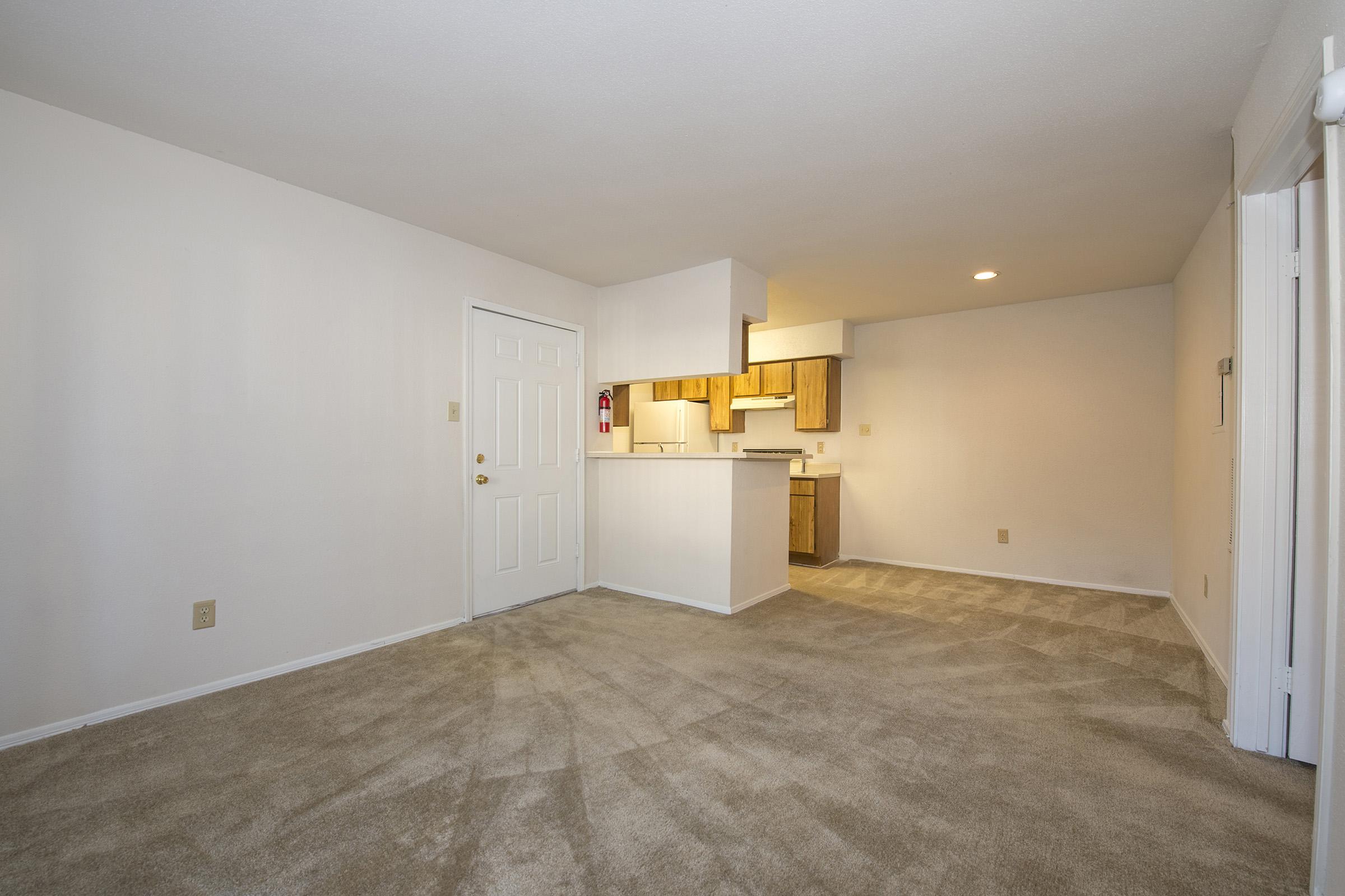 Empty room with beige carpet and white walls. A door is visible on the left side, leading outside. In the background, there is a small kitchen area with wooden cabinets. Soft natural light is coming from an overhead ceiling light, creating a bright and open atmosphere.