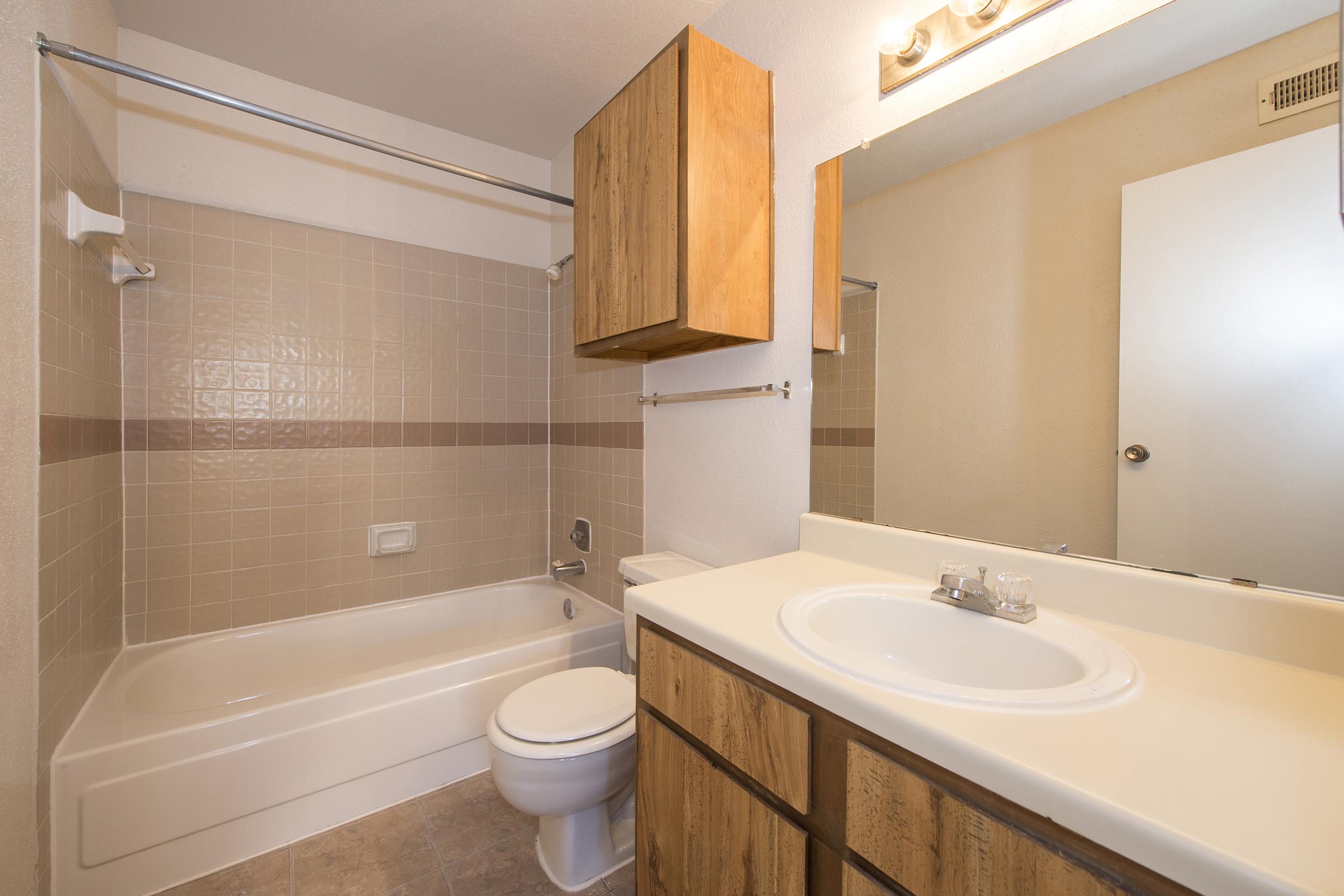 A clean and simple bathroom featuring a white bathtub with a shower, a wooden vanity with a sink, and a mirror above it. The walls are tiled in light beige with a decorative stripe, and there is a light fixture above the mirror. Storage cabinets are mounted above the toilet and vanity.