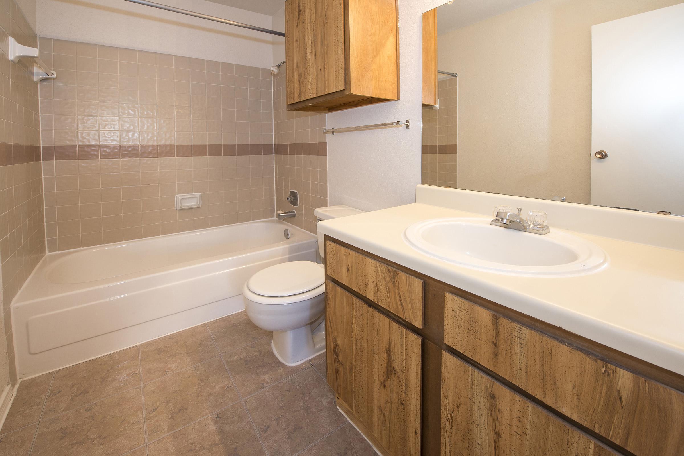 A clean and simple bathroom featuring a light-colored tiled shower/bathtub area, a modern toilet, and a brown wooden vanity with a white sink. A large mirror reflects the surroundings, and the walls are painted in neutral tones. The floor is tiled with a light pattern, enhancing the bright and spacious feel.