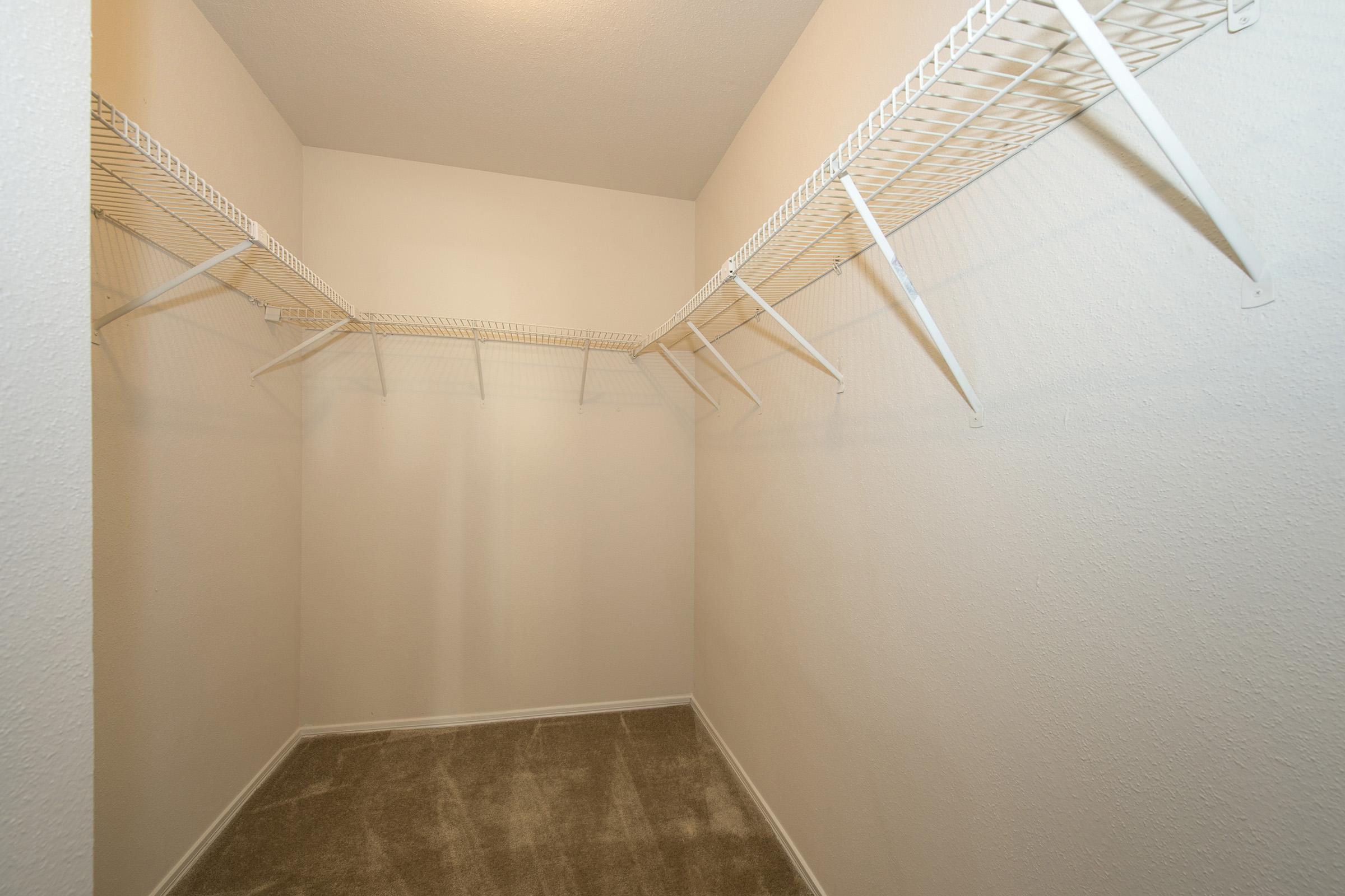 A spacious, empty closet featuring wire shelving attached to the walls. The walls are painted a neutral color, and the floor is covered with beige carpet. The overall appearance is bright and simple, with ample room for storage.