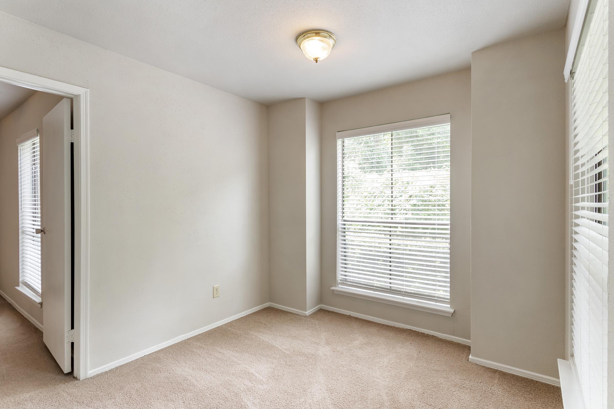 Empty room with light beige walls and carpet. A single window with white blinds allows natural light to enter. A ceiling light provides illumination, and there is a partially open door leading to another space. The overall atmosphere is bright and airy, creating a minimalist feel.