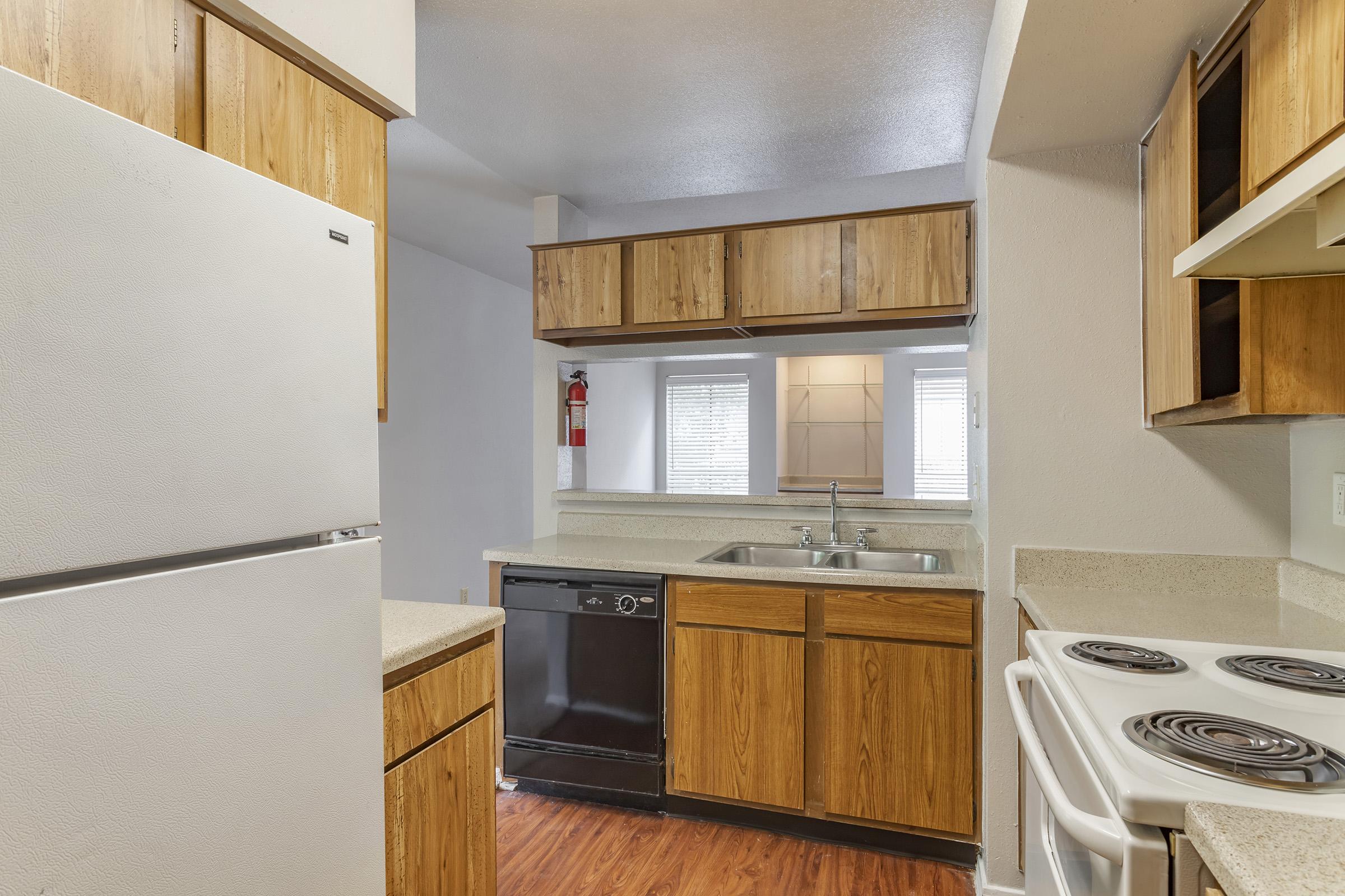 A modern kitchen featuring wooden cabinets, a sink, and countertop space. Appliances include a white refrigerator, a black dishwasher, and a white stove with burners. The kitchen has natural light coming in from a nearby window, along with a neutral color palette and laminate flooring.