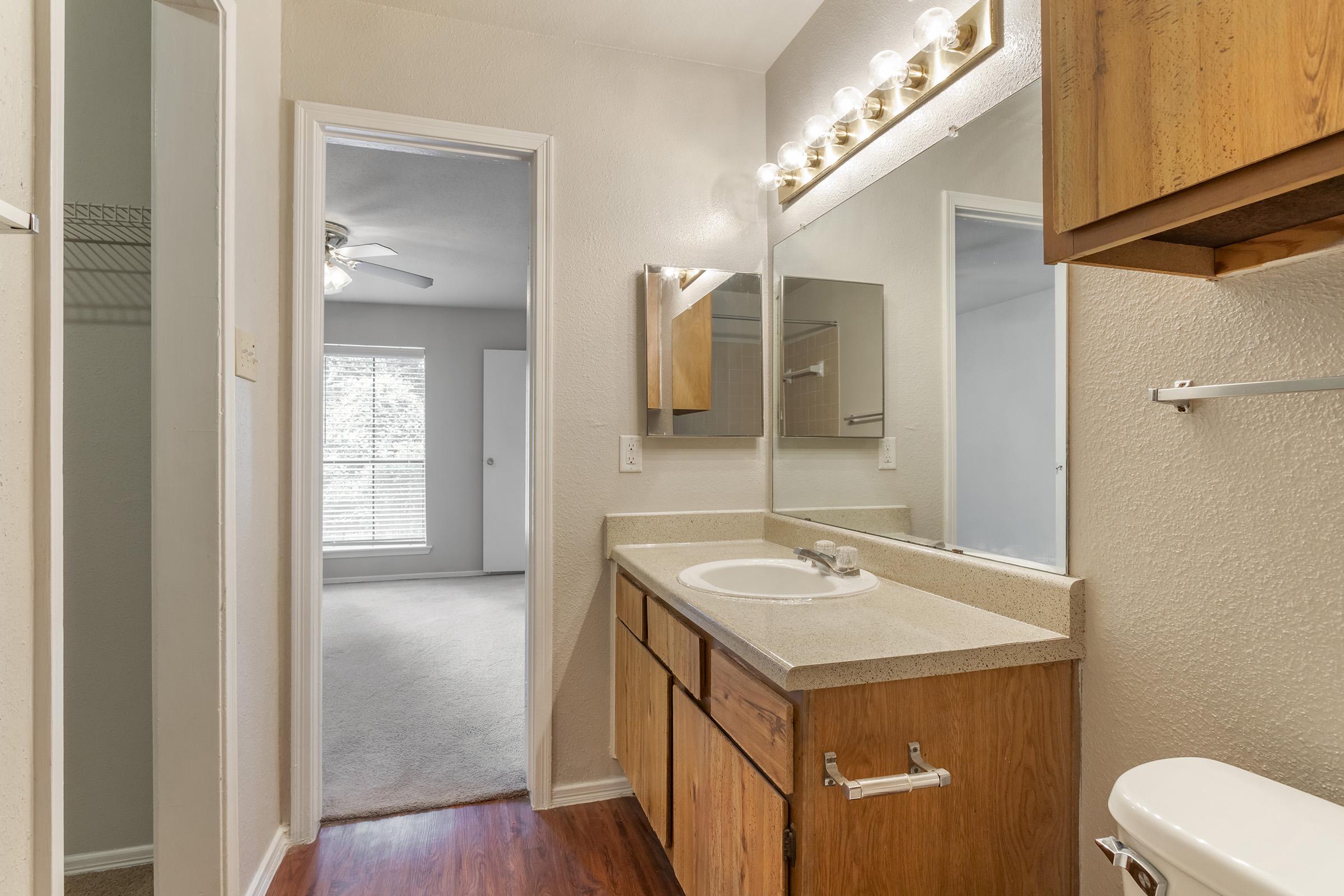 A well-lit bathroom featuring a single sink vanity with a large mirror, wooden cabinets, and a countertop. In the background, a spacious room with windows displaying natural light and neutral-colored walls can be seen. The flooring combines tiles and carpet, creating a cozy atmosphere.