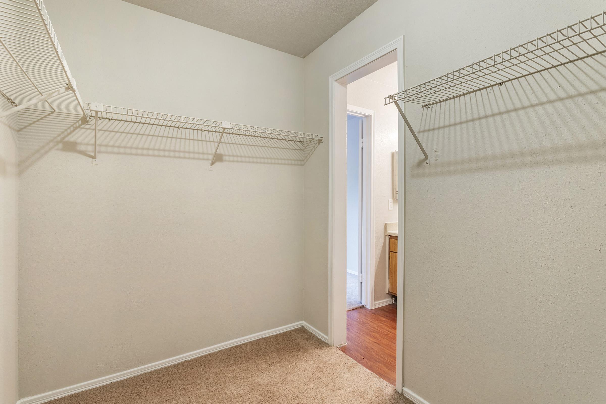 Empty closet with white wire shelving on two walls, light-colored walls, and carpeted floor. A doorway on the left leads to a bathroom or another room, revealing a hint of a light-colored floor and a wall. The overall space is well-lit and tidy, appearing ready for organization.