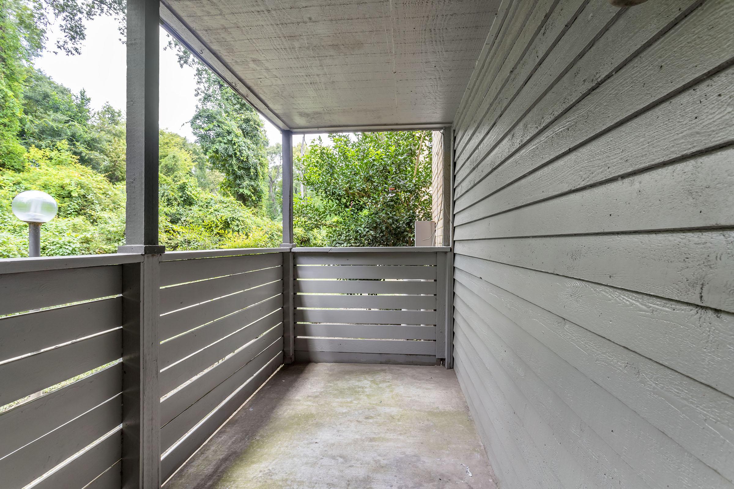 A covered balcony with gray wooden railing and a concrete floor. There are lush green trees and bushes visible in the background, creating a natural, serene atmosphere. A round outdoor light fixture is situated on the left side of the balcony. The walls are painted in a light gray shade.