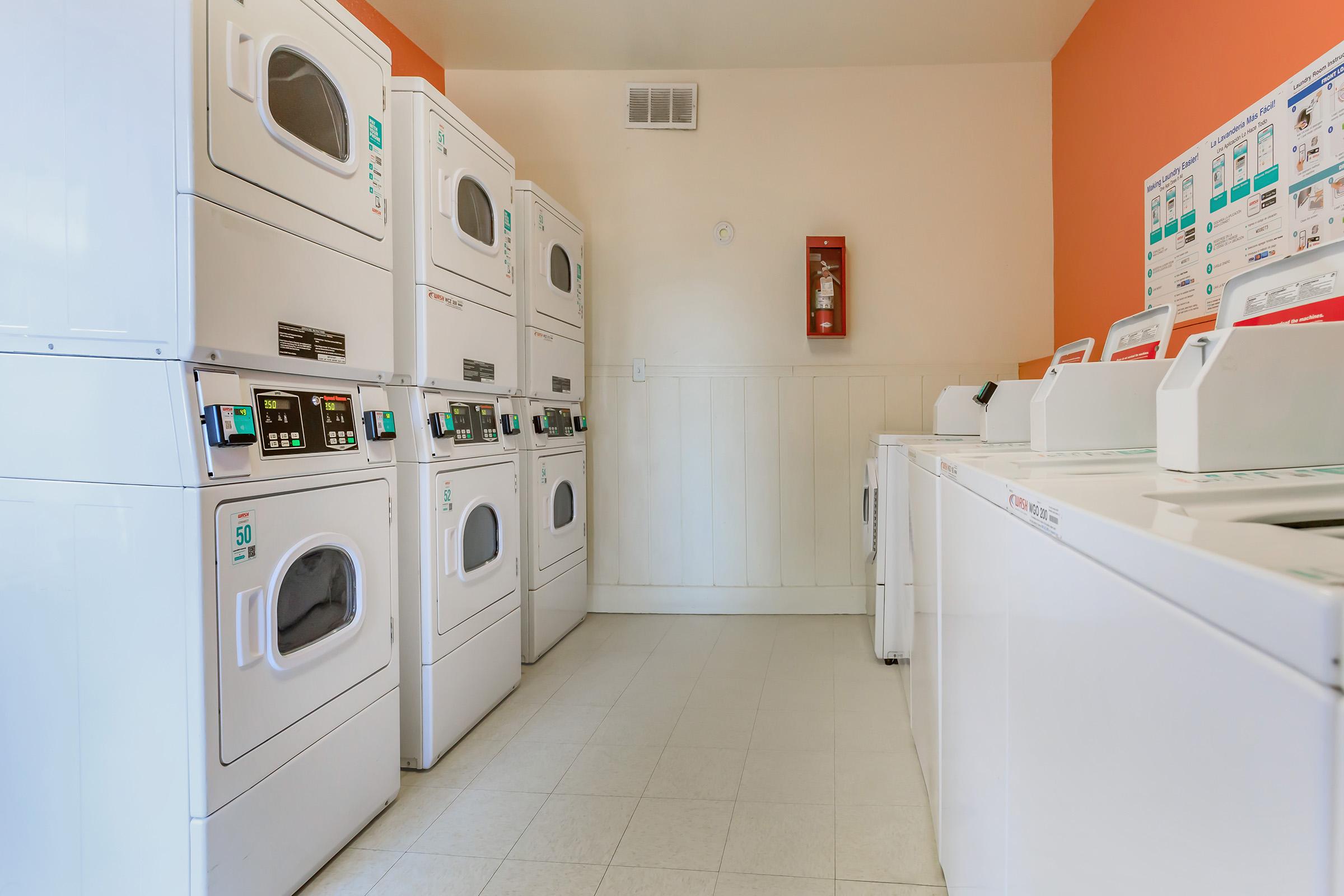 A clean laundry room featuring several stacked white washers and dryers along the walls. The room has orange accents, a fire extinguisher mounted on the wall, and informational posters on the right side. The floor is tiled in light colors, creating a bright and organized space for laundry.