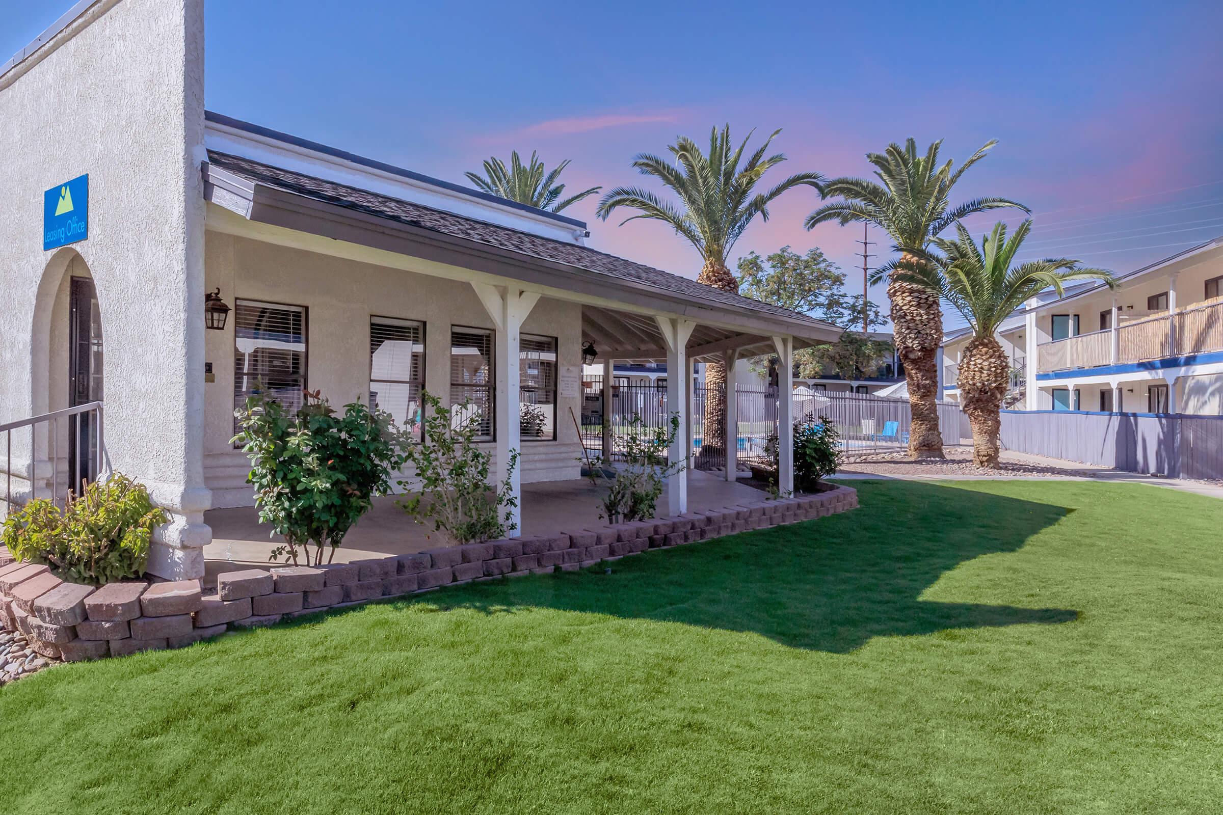 A modern home exterior featuring a patio with white pillars and a well-maintained lawn. Palm trees surround the backyard, and there is a clear blue sky. The house is painted in light colors and has windows with elegant trim, creating a welcoming outdoor space.