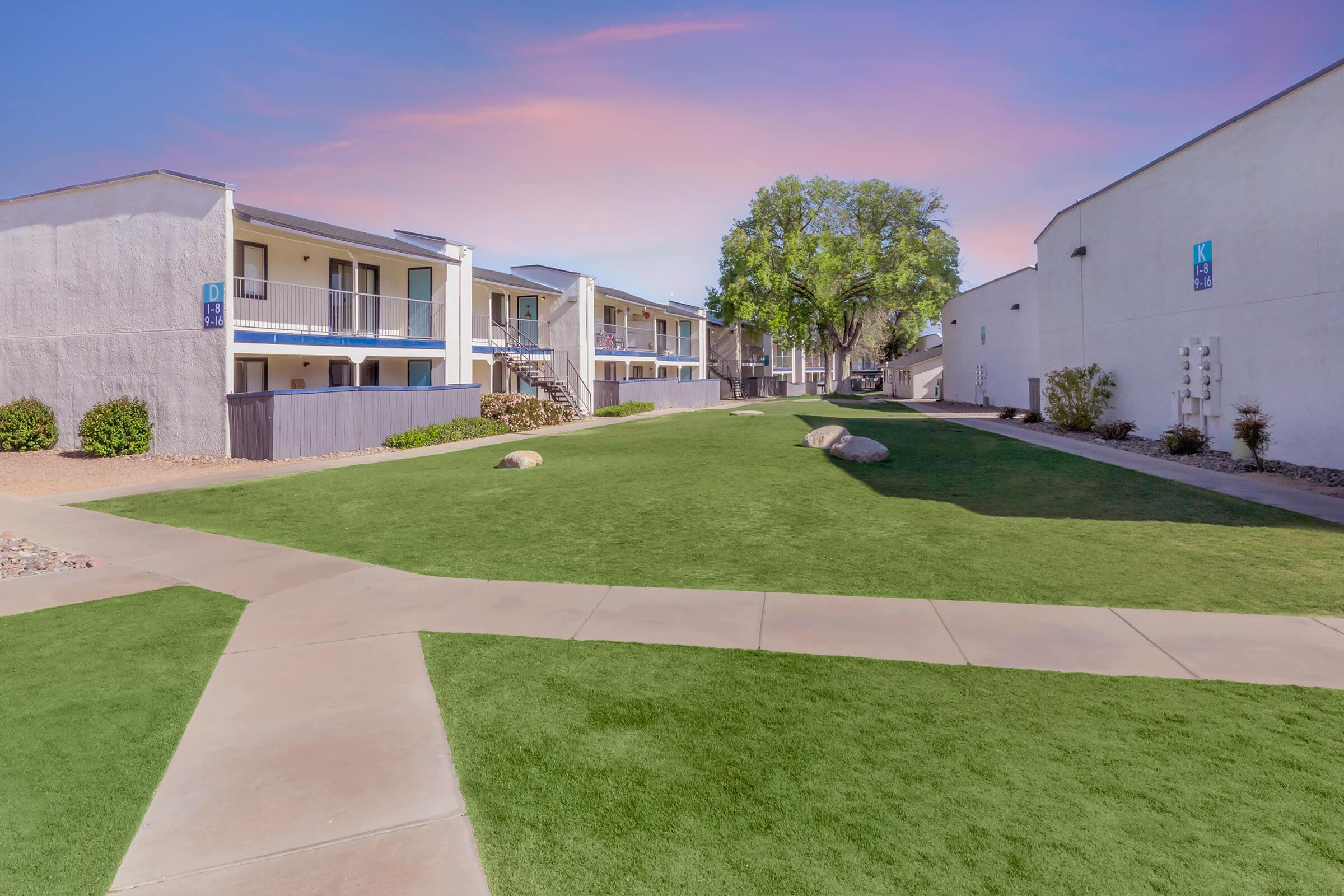 Green lawn with small rocks, bordered by two apartment buildings with balconies. A sidewalk runs through the lawn, creating pathways that intersect. In the background, a tree adds to the landscaping against a pastel-colored sky.
