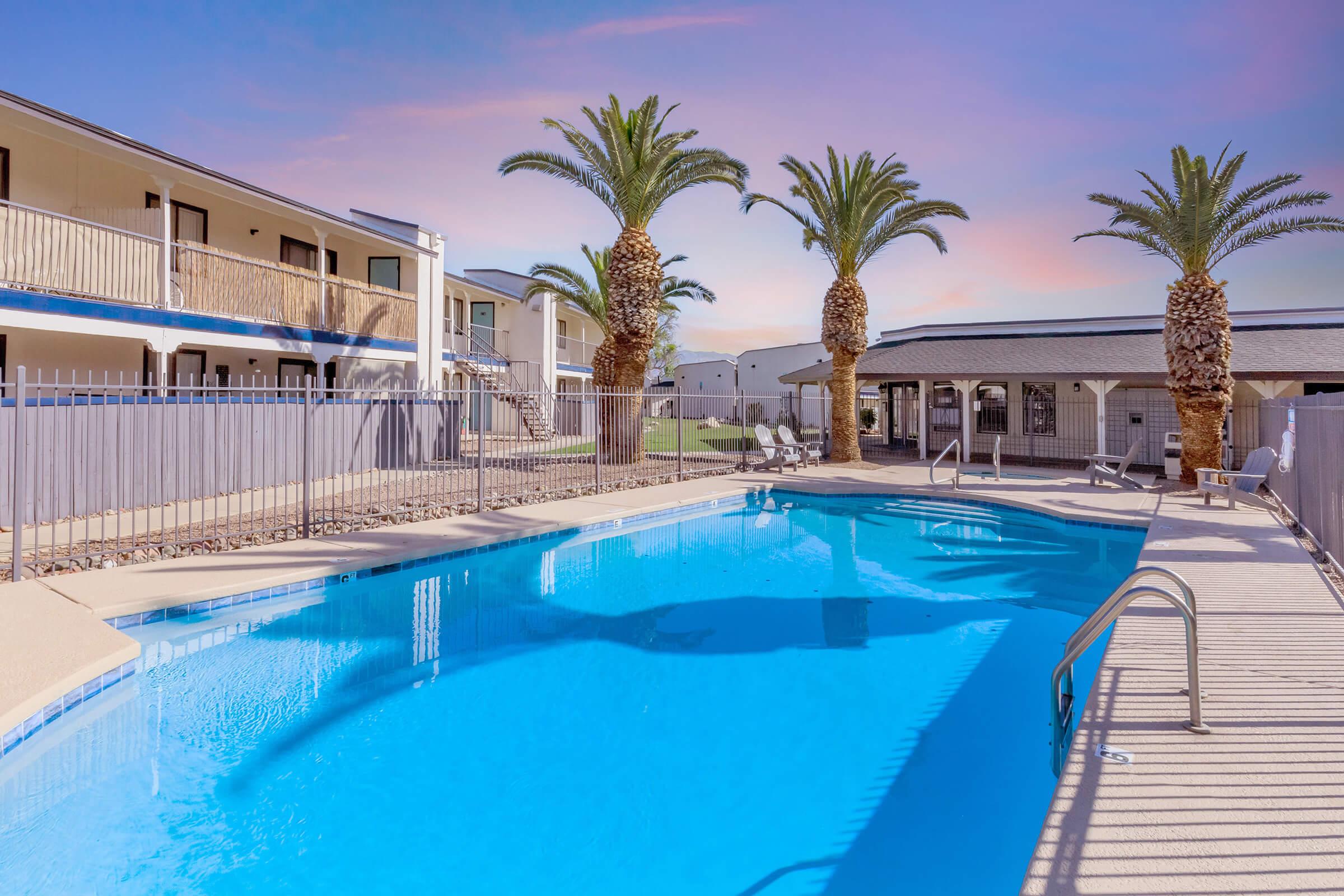 A clear blue swimming pool surrounded by palm trees, with lounge chairs and a wooden deck. In the background, two-story buildings with balconies and a fenced area complete the tranquil setting, under a pastel sky at dusk.