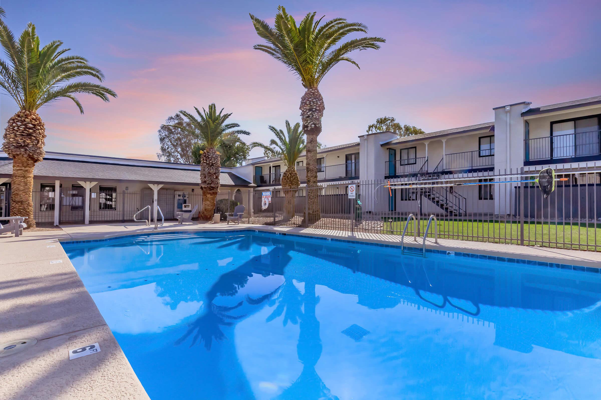 A clear swimming pool surrounded by palm trees, with a railing and lounge chairs nearby. In the background, a building with multiple rooms is visible, and the sky displays a colorful sunset. The atmosphere is inviting and relaxing.