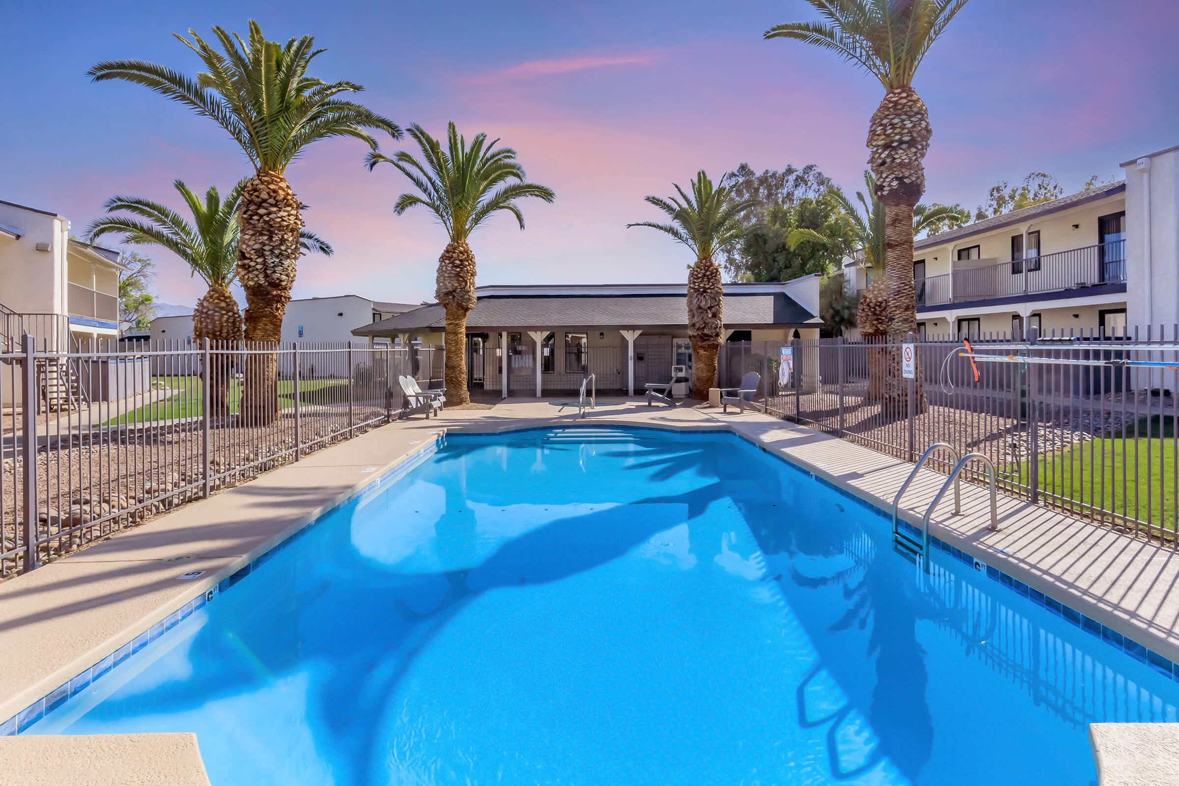 A clear blue swimming pool surrounded by palm trees, with a fenced area. In the background, there is a building featuring a covered patio space and additional structures. The sky is a gradient of pink and purple, suggesting a sunset atmosphere.