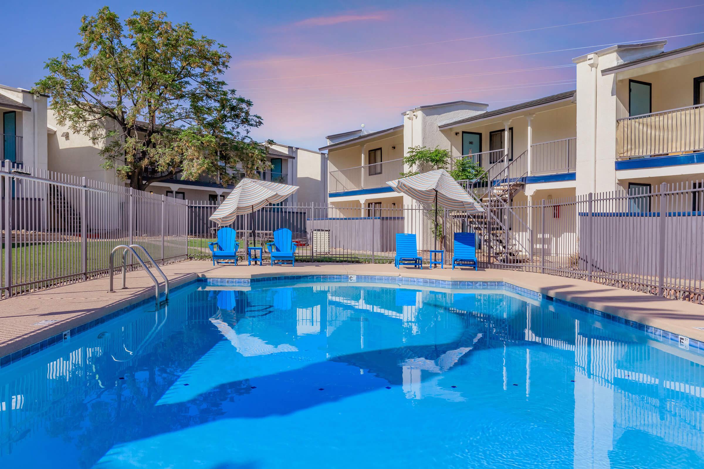 A swimming pool surrounded by lounge chairs and shaded areas, located in a courtyard with two-story buildings in the background. The pool water is clear, reflecting the sky. Trees and greenery are visible around the pool area.