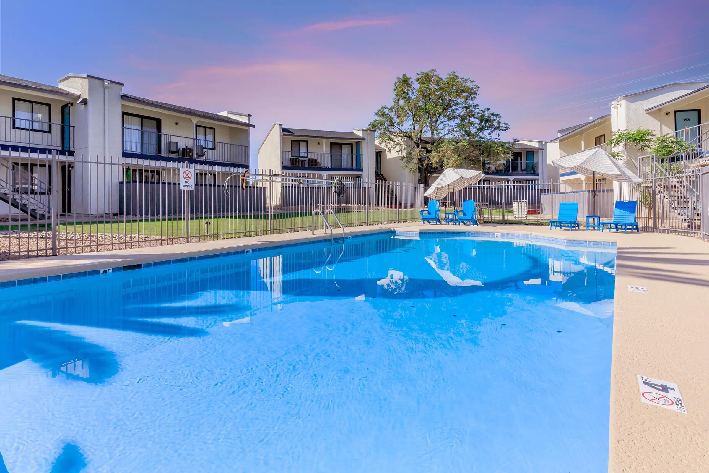 A sparkling blue swimming pool surrounded by a gated fence, with lounge chairs nearby. In the background, several apartment buildings are visible, along with green grass and a tree, under a colorful sky.