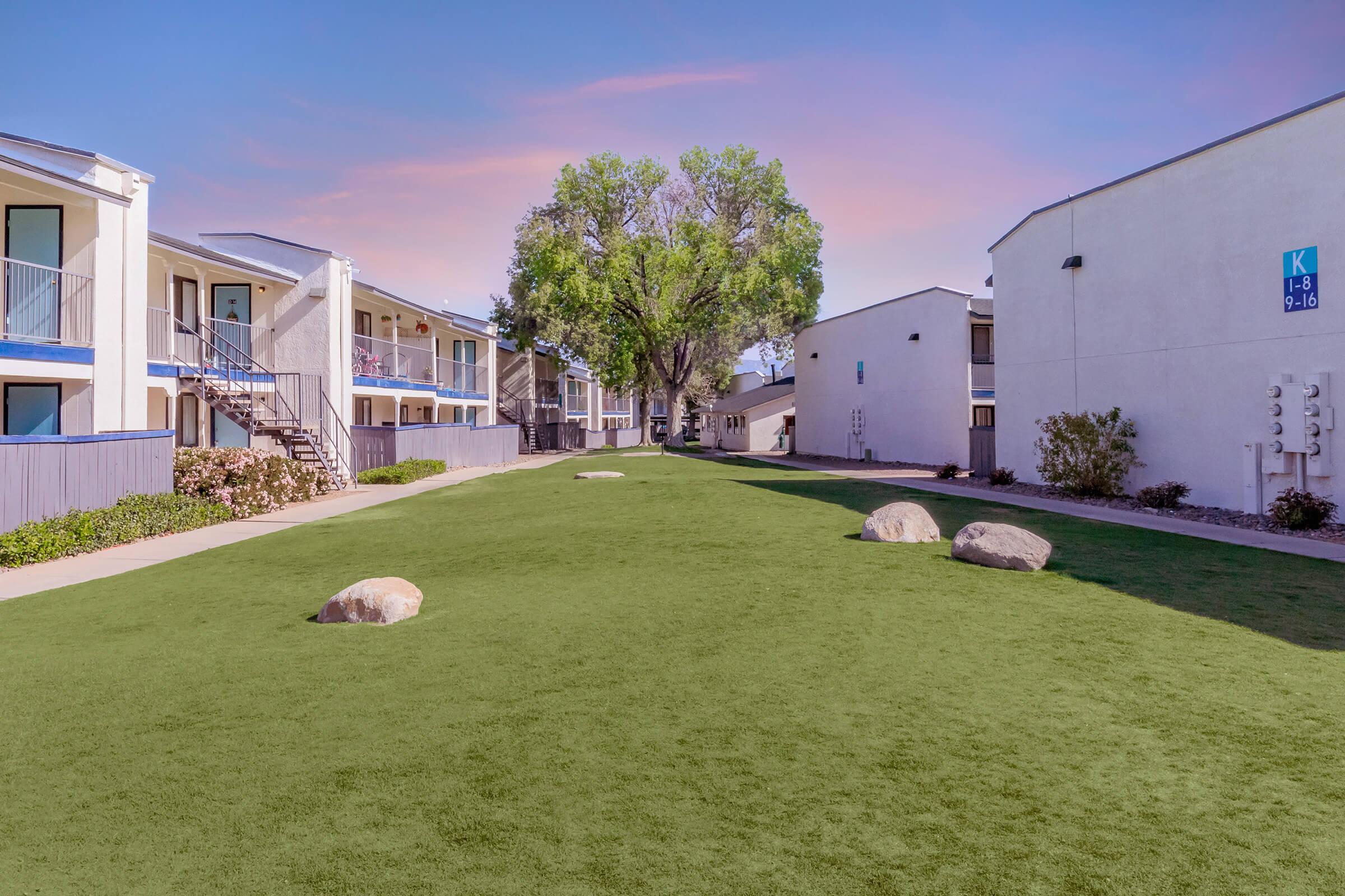 A well-maintained courtyard featuring green artificial turf, flanked by two multi-story apartment buildings. The space includes a few large rocks and is surrounded by shrubs and trees under a clear blue sky with a hint of colorful sunset hues.