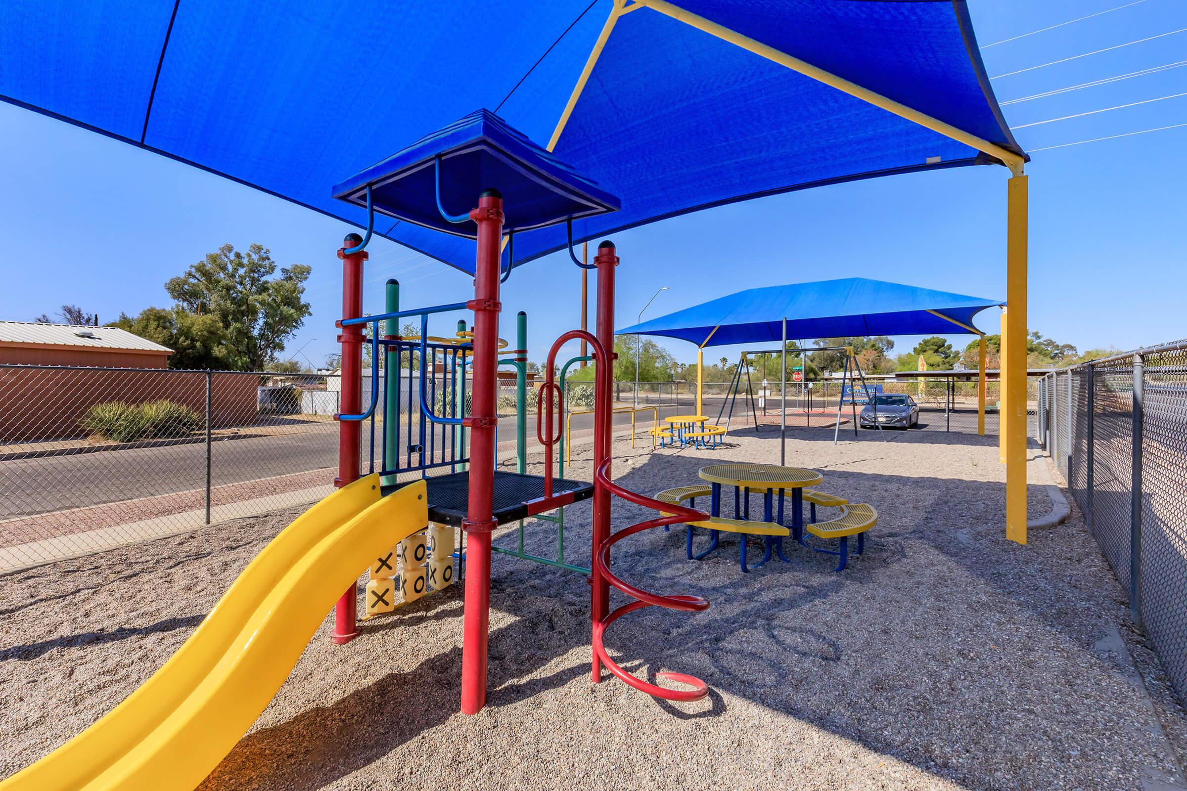 Playground featuring a yellow slide, climbing structure with colorful poles under a large blue shade sail. Nearby, there are yellow picnic tables and a fenced area. The playground is situated in an open space with clear skies and some trees in the background.
