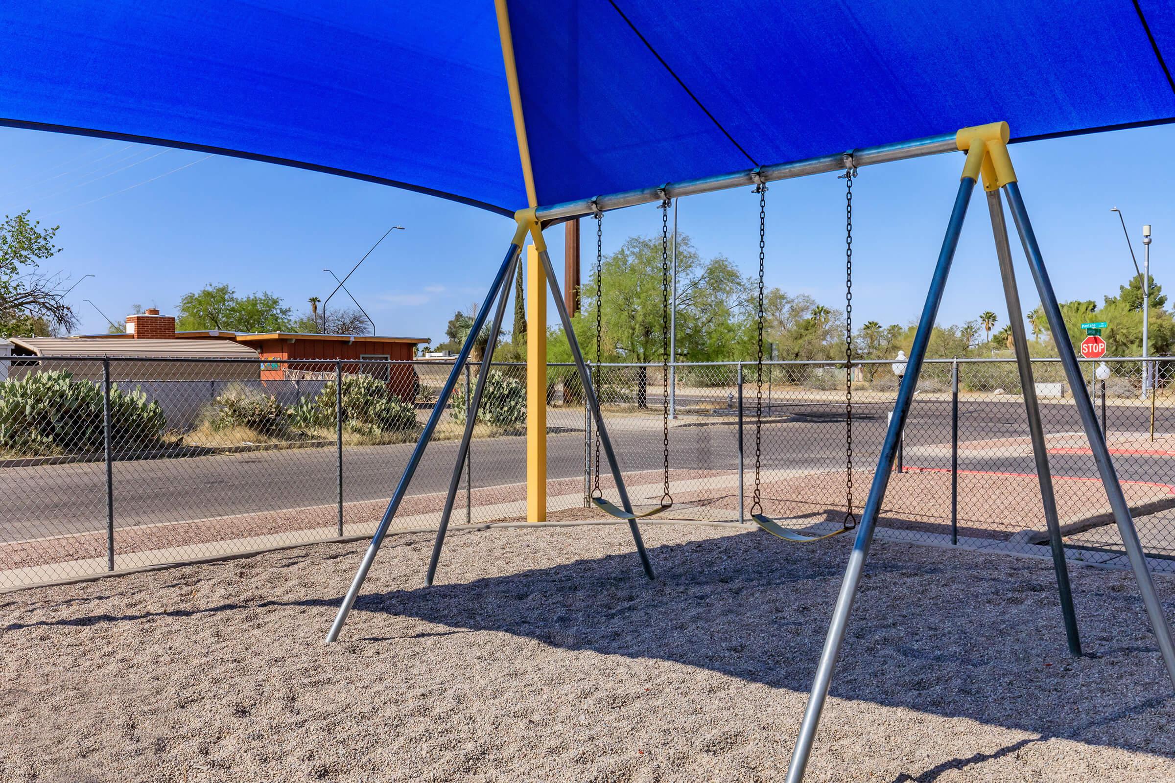 A playground featuring two swings under a blue shade structure, surrounded by gravel. In the background, there are trees and a residential area behind a fence. The setting is sunny and open with a clear blue sky.