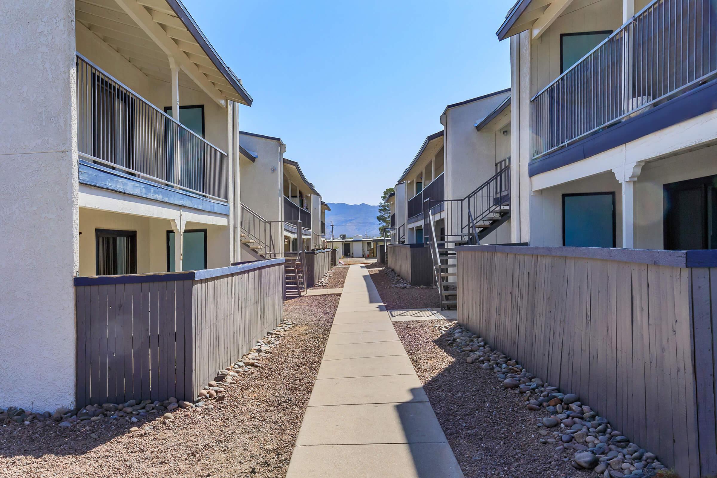 A walkway between two rows of buildings, leading to a distant view of mountains. The buildings are white with balconies, and the pathway is lined with gravel and wooden fences. Clear blue sky above.