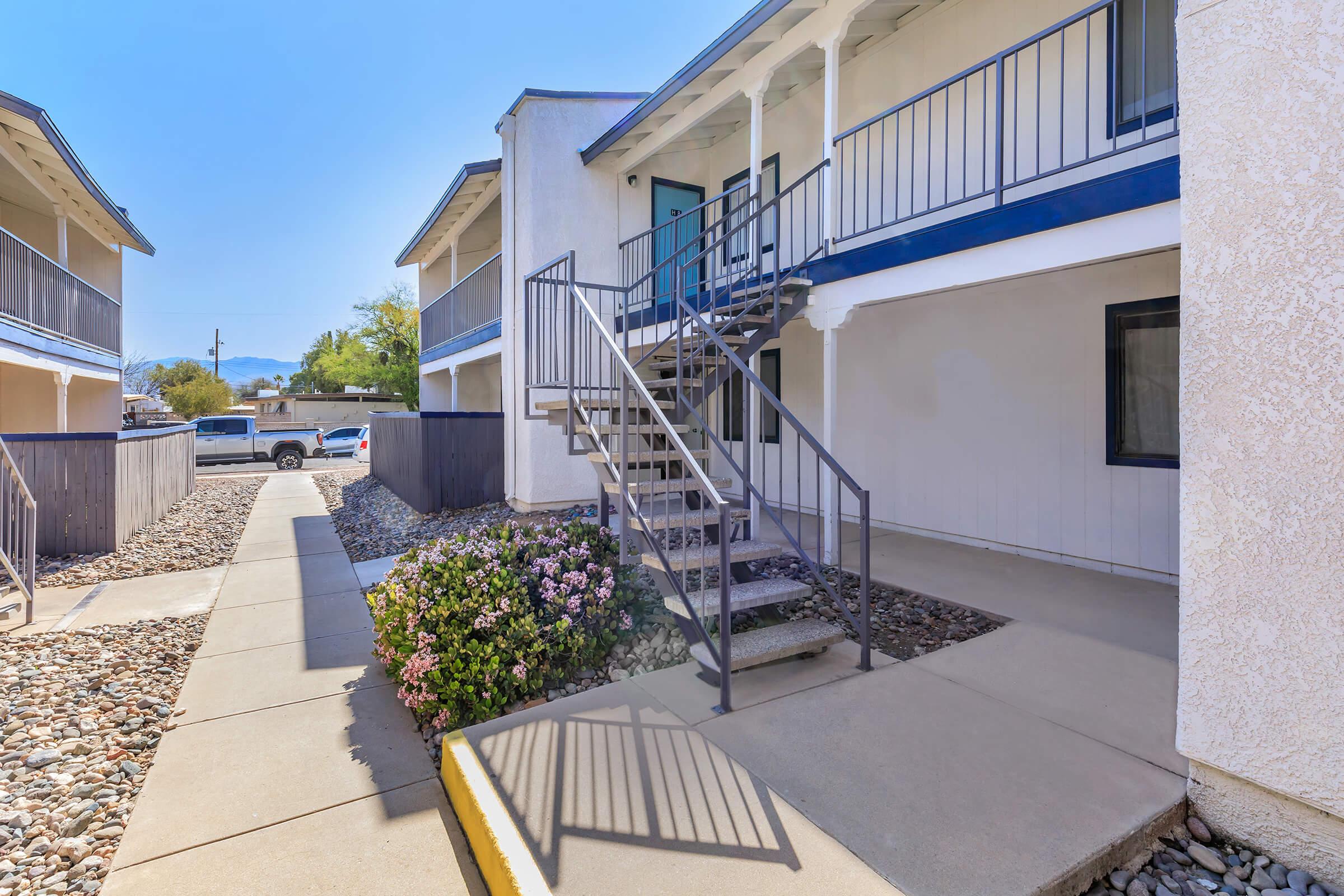 Exterior view of a two-story apartment building featuring a set of metal stairs leading to the second floor. The pathway is lined with gravel and small bushes, and there are parking spaces visible in the background under a clear blue sky.