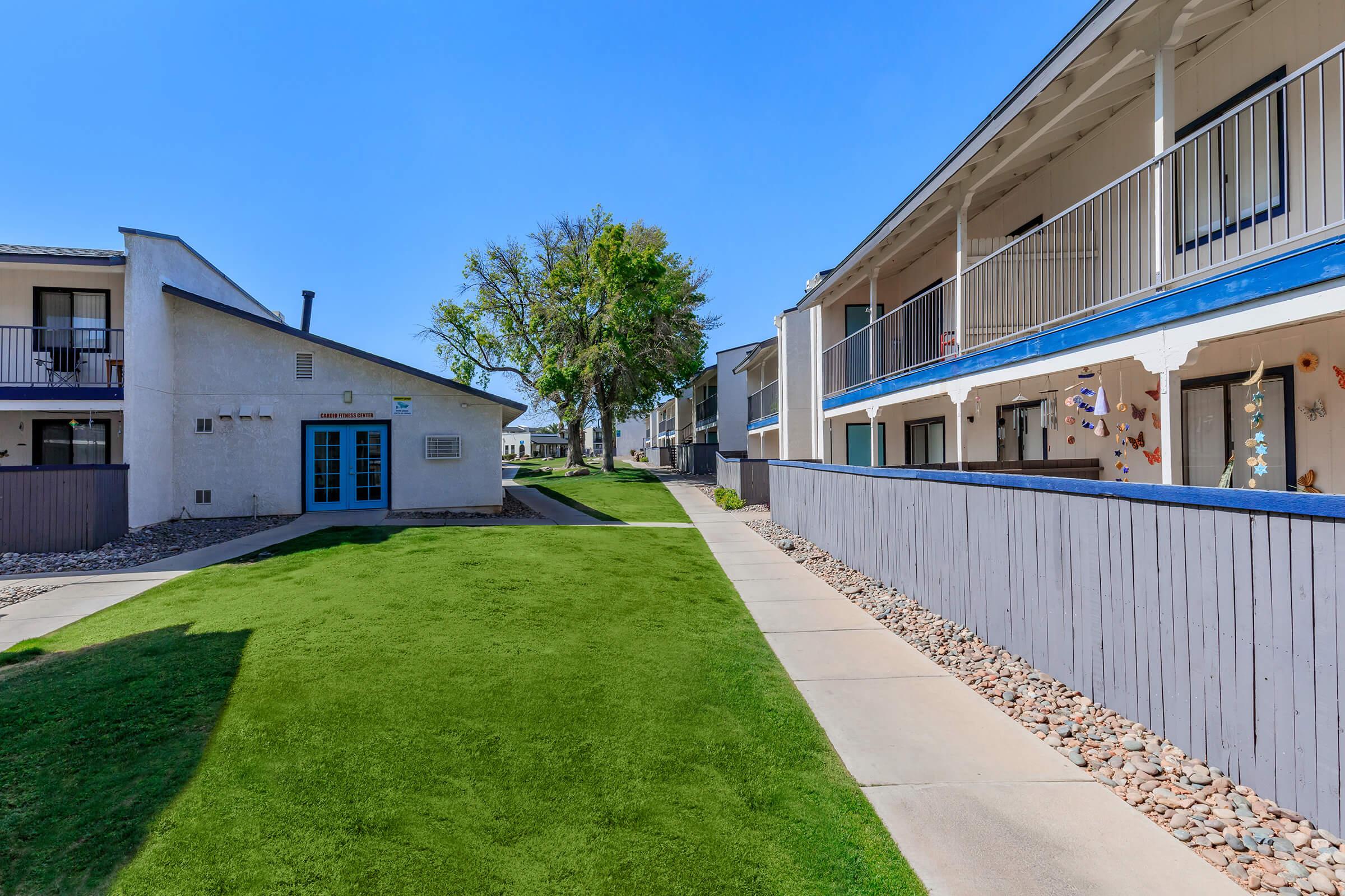 A well-maintained residential area featuring two building structures on either side of a grassy pathway. One side has a blue door and a tree, while the other has balconies and colorful decorations. The sky is clear and blue, suggesting a sunny day.