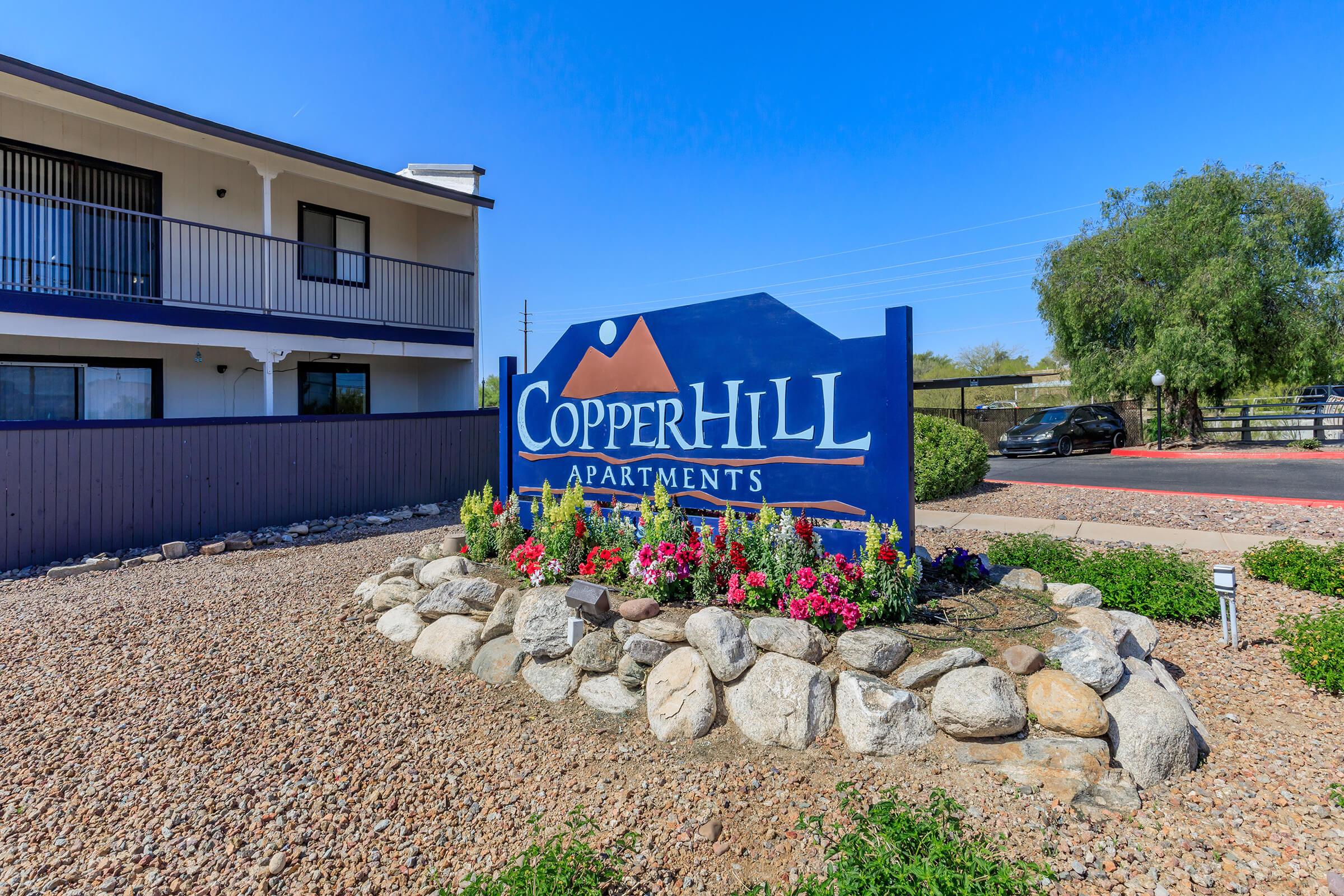 Sign for Copperhill Apartments surrounded by flowers and rocks, with a clear blue sky overhead. The apartment building is visible in the background, featuring balconies and a modern design.