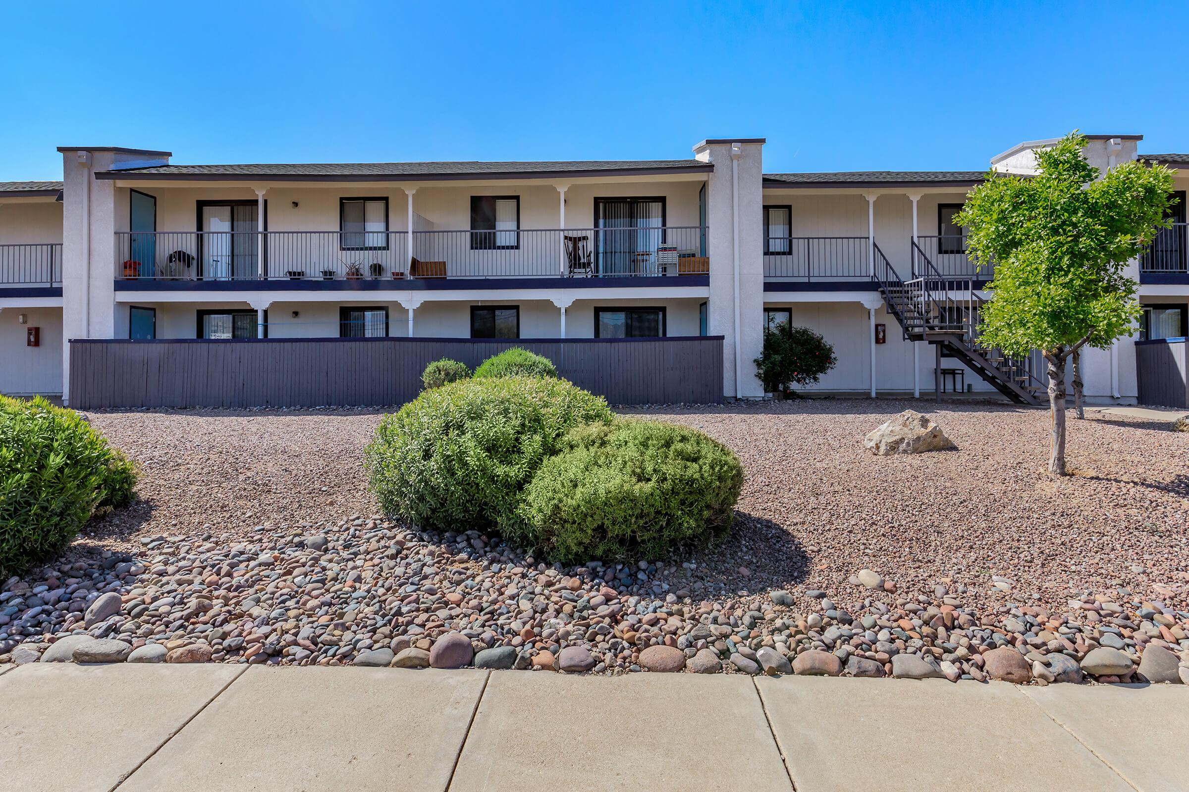 Two-story hotel building featuring multiple balconies with black railings. The ground level has a gravel landscape with small shrubs and a decorative rock. A small tree is visible to the right, under a clear blue sky. The overall setting appears bright and welcoming.