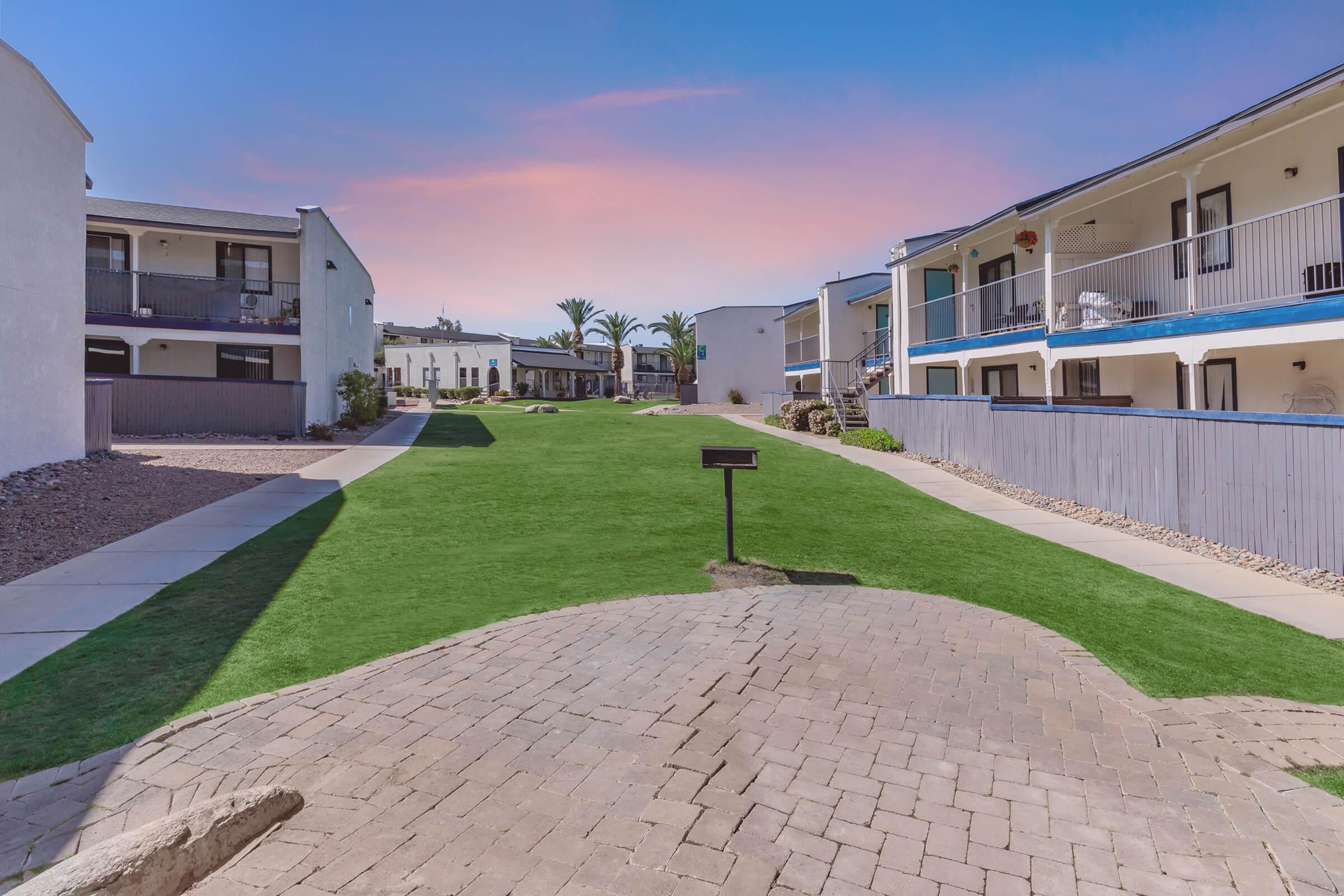 A landscaped area between two buildings, featuring green grass and a brick pathway. The scene includes palm trees in the background and a blue sky at sunset, creating a serene atmosphere. The buildings have balconies and a contemporary design, contributing to a welcoming outdoor space.