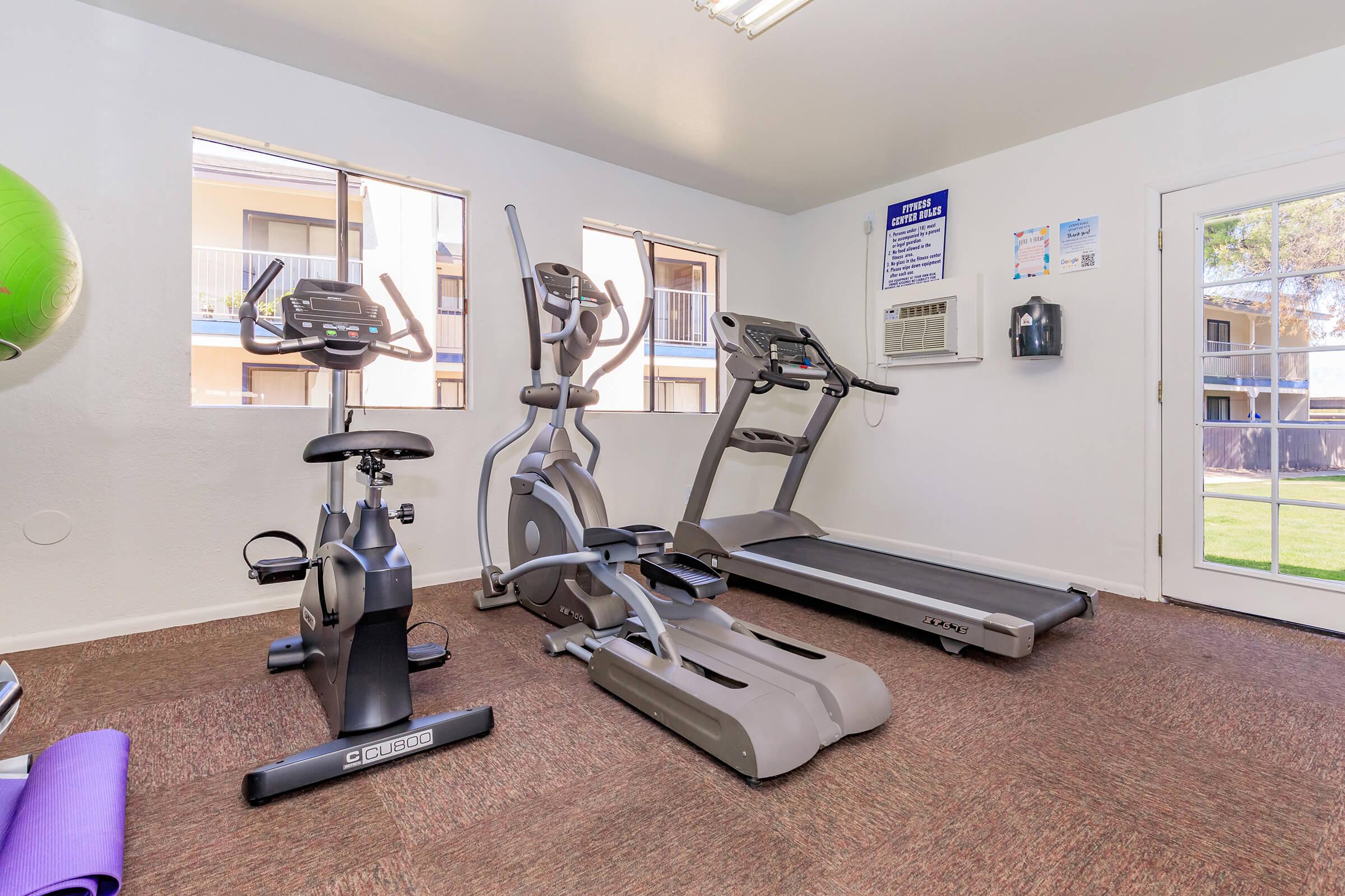 A well-lit gym with exercise equipment, including a stationary bike, elliptical machine, and treadmill. There are windows providing natural light, wall-mounted air conditioning unit, and a door leading outside. The flooring is carpeted, and exercise mats are placed on the floor.