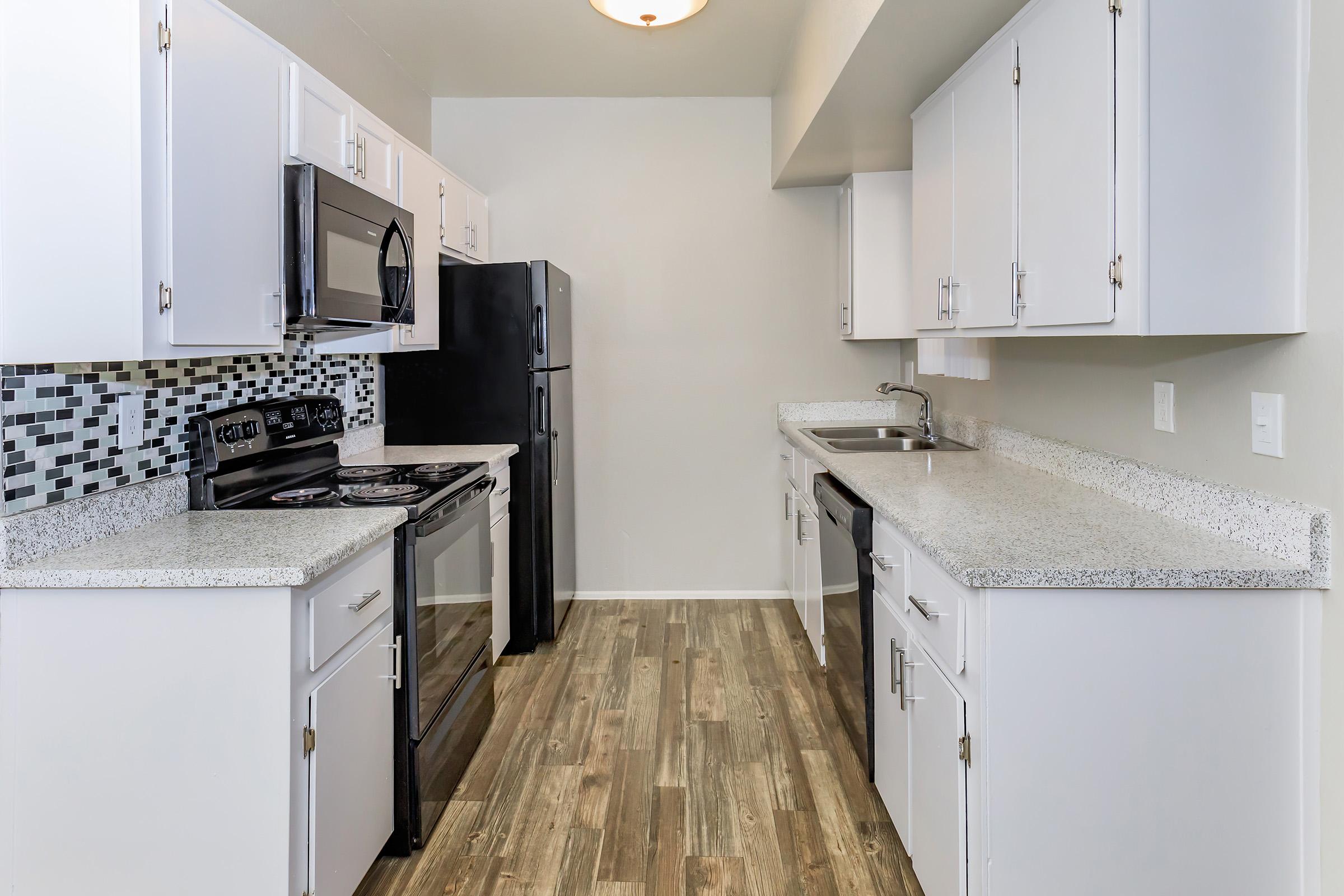 Modern kitchen featuring white cabinetry, granite countertops, and stainless steel appliances, including a microwave, stove, and refrigerator. The backsplash consists of black and gray tiles, and the floor is styled with wood-like vinyl planks. Well-lit with a ceiling fixture.
