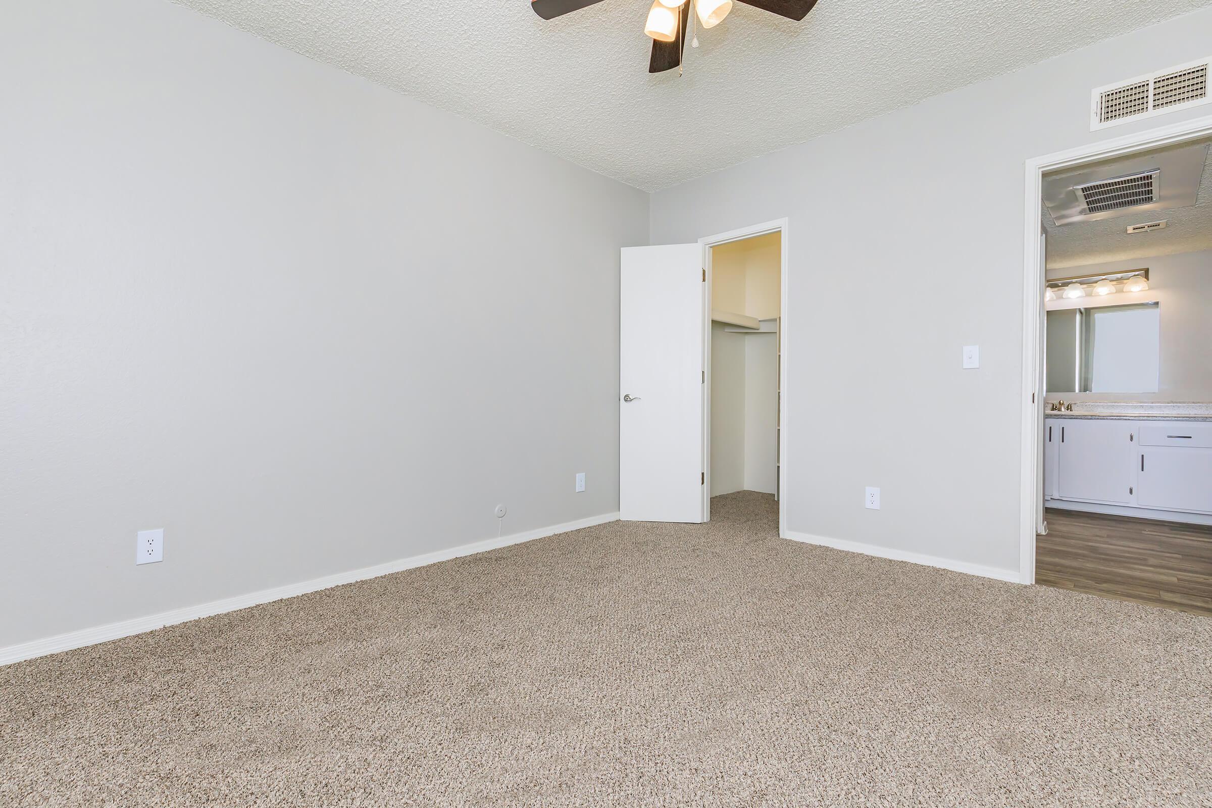 A spacious, empty room with beige carpet, light gray walls, and a ceiling fan. A doorway leads to a closet, while another doorway opens to a bathroom area. The room is well-lit, featuring natural light and a clean, minimalistic design.