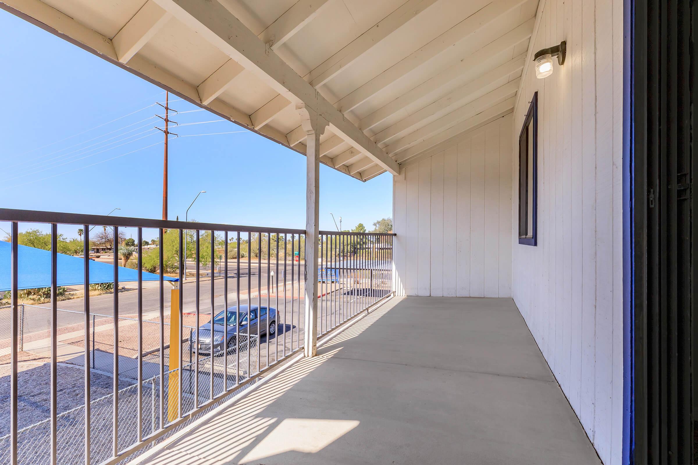 View of a balcony with a concrete floor, featuring a railing. The scene shows a clear blue sky, utility poles in the distance, and a neighborhood below. The balcony is part of a white building with a slanted roof. The area appears bright and spacious, suitable for outdoor relaxation.