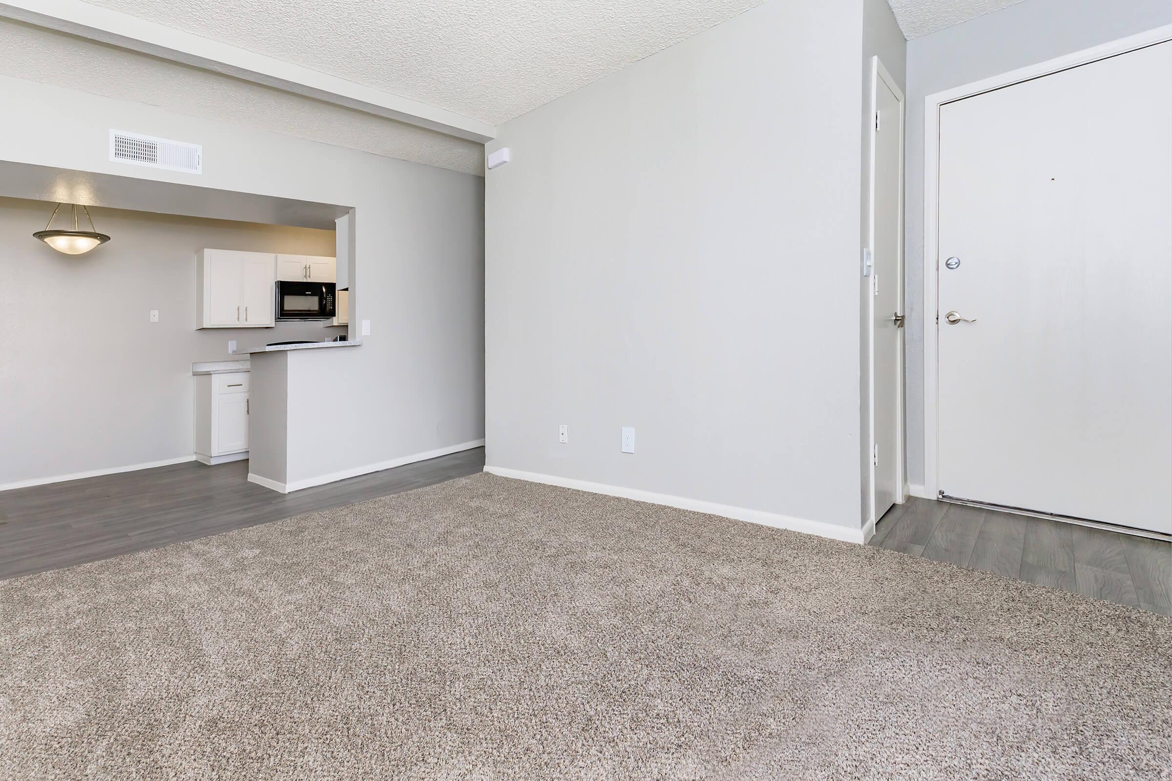 A spacious, well-lit living area with light-colored walls and gray carpet. There’s an open kitchen in the background with white cabinets and modern appliances. A front door is visible on the right side, and the layout appears welcoming and cozy. Ideal for a comfortable living space.