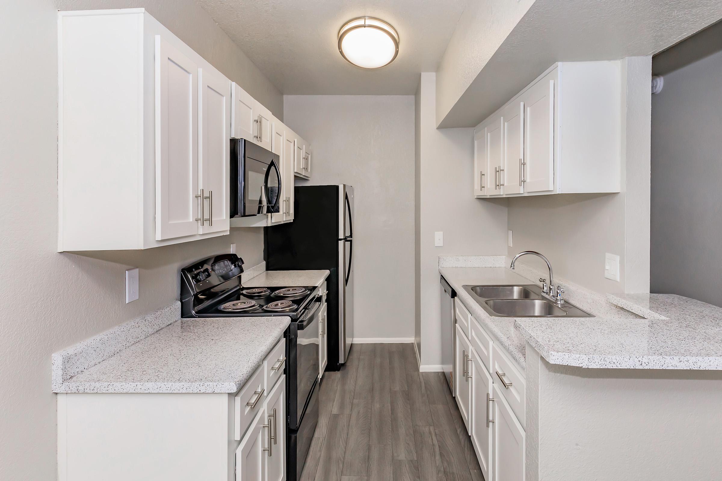 Modern kitchen with white cabinets, a black oven and refrigerator, and gray countertops. The space has a light gray wall color and wood-like flooring, with a single basin sink and a ceiling light fixture. The kitchen is designed for efficiency with ample storage and counter space.