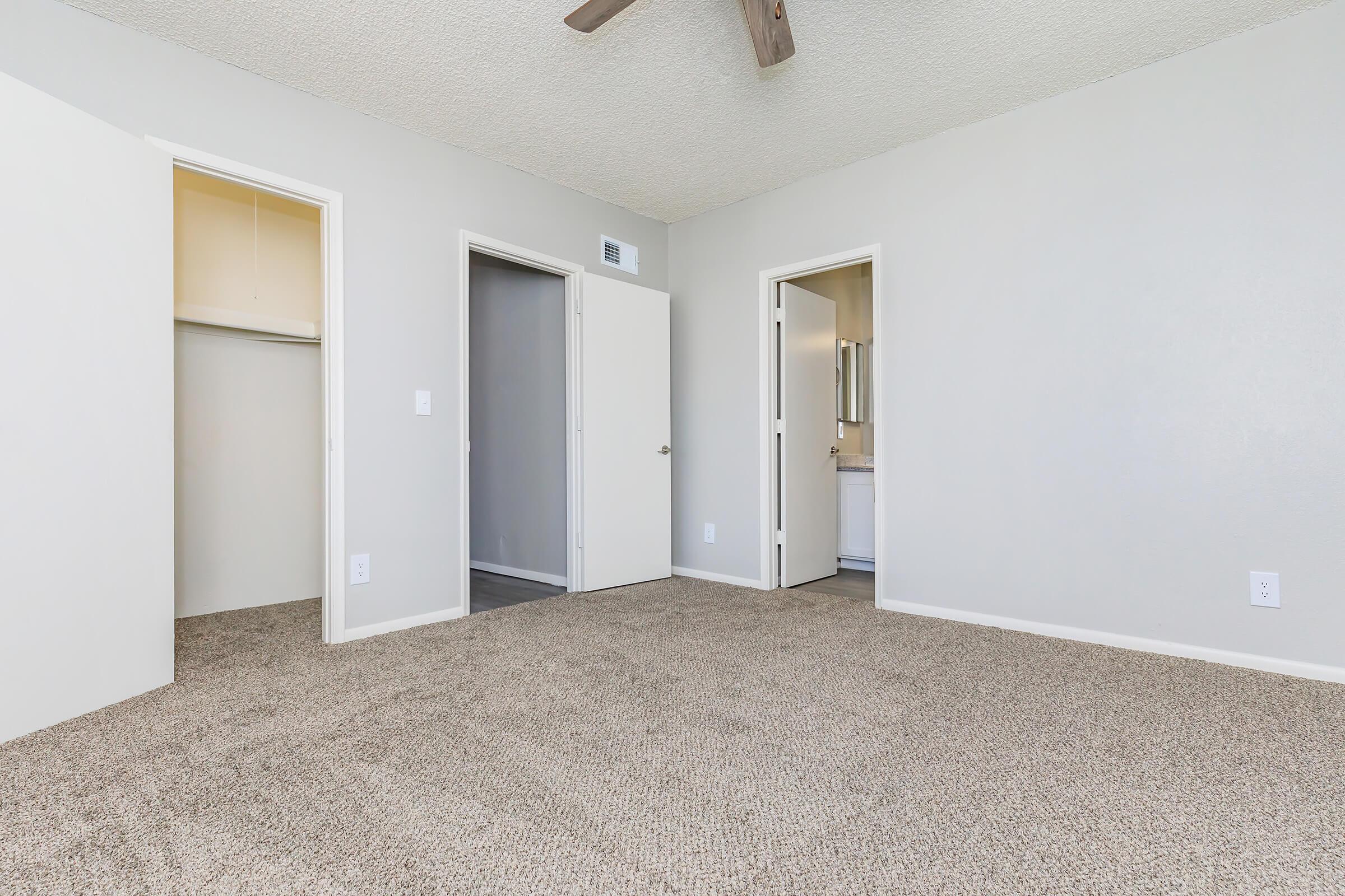 A spacious room featuring beige carpet, textured ceiling, and three doorways leading to a closet and two separate areas. The walls are painted in a light gray color, and there is a ceiling fan with wooden blades. Natural light is minimal, creating a calm and neutral atmosphere.
