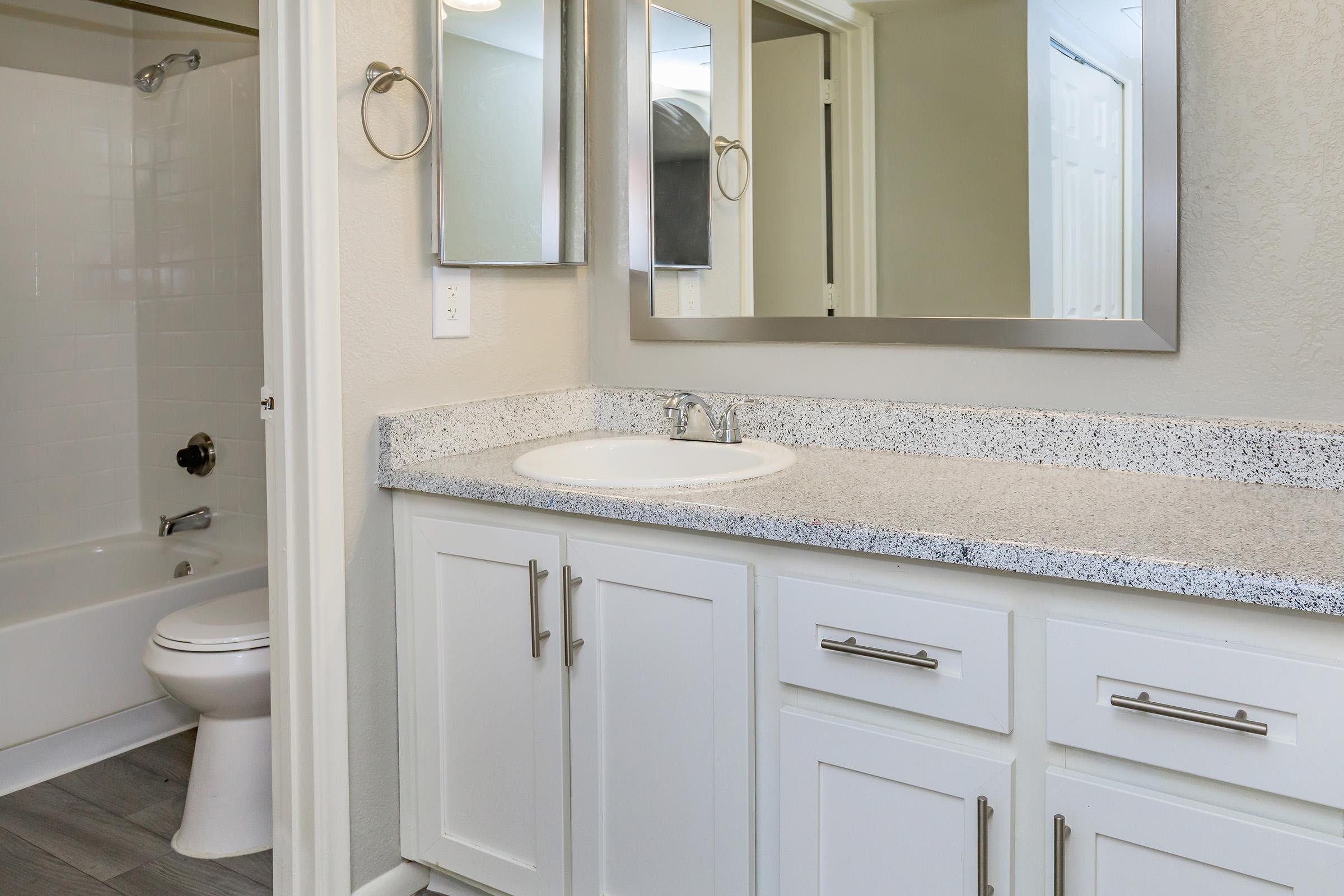 A modern bathroom featuring a white granite countertop with a sink, a large mirror above, and a cabinet with silver handles. A white bathtub is visible to the left, with light beige walls and grey flooring, creating a clean and elegant look.
