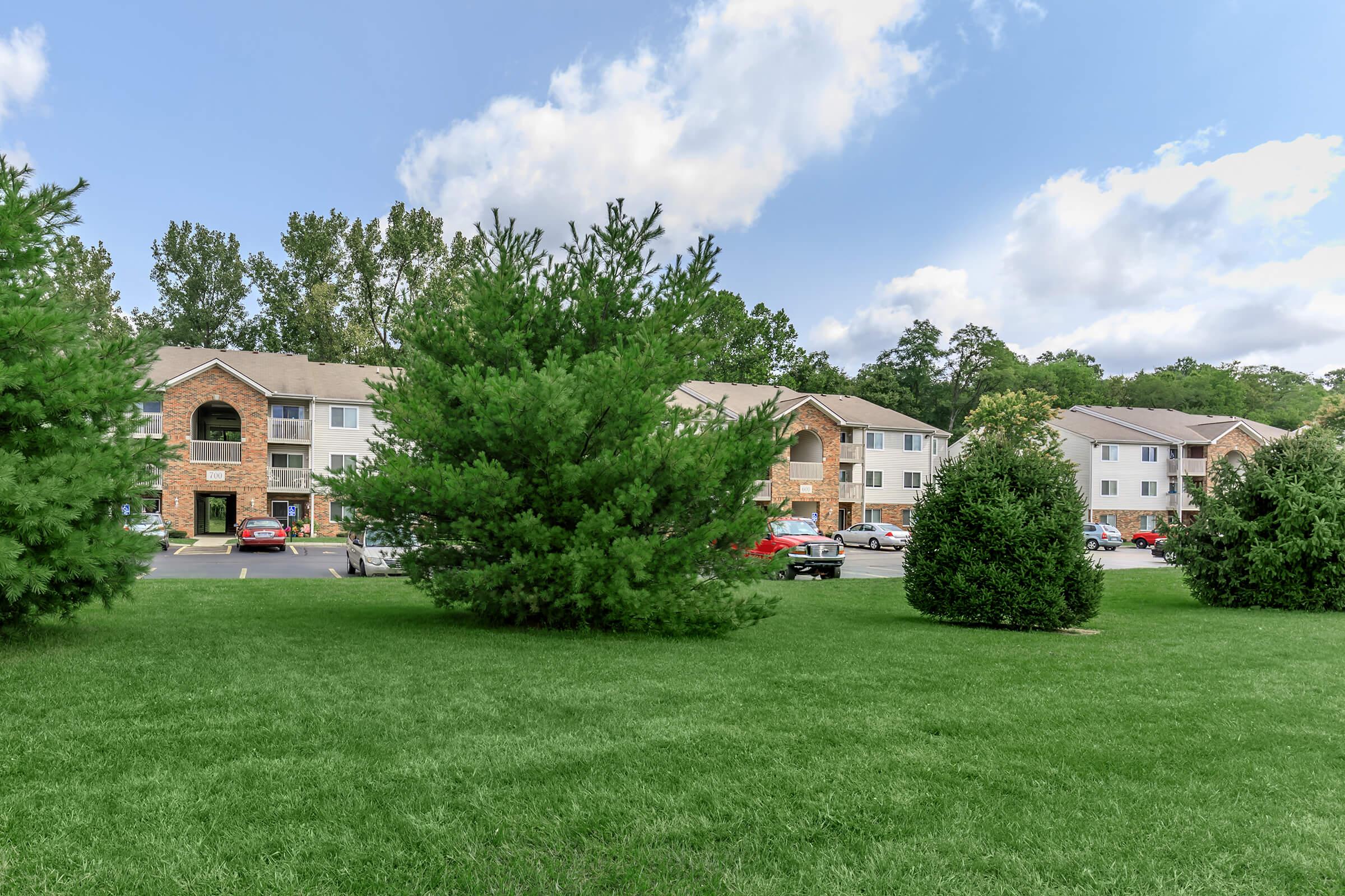 View of a well-maintained residential complex featuring multiple apartment buildings. In the foreground, there is a green lawn with bushes, while in the background, trees line the property, and parked cars are visible in the lot. The sky is partly cloudy, creating a pleasant atmosphere.