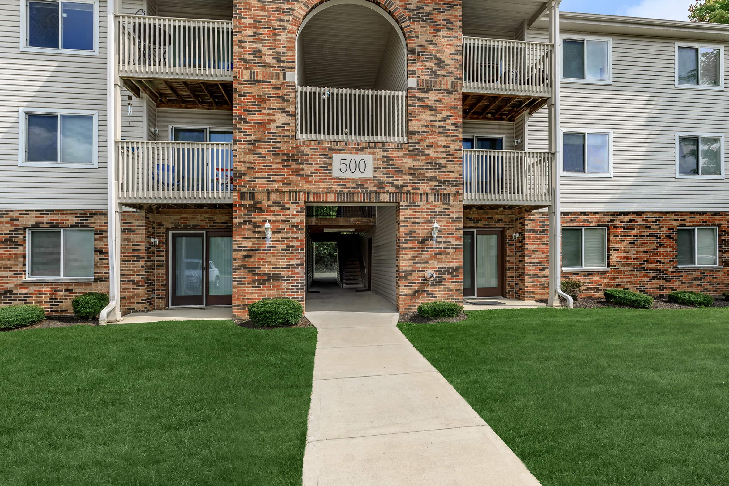 Exterior view of an apartment building featuring a brick facade, multiple balconies, and a central entrance. The pathway leads to the entrance numbered 500, with well-maintained grass and shrubs surrounding the area.
